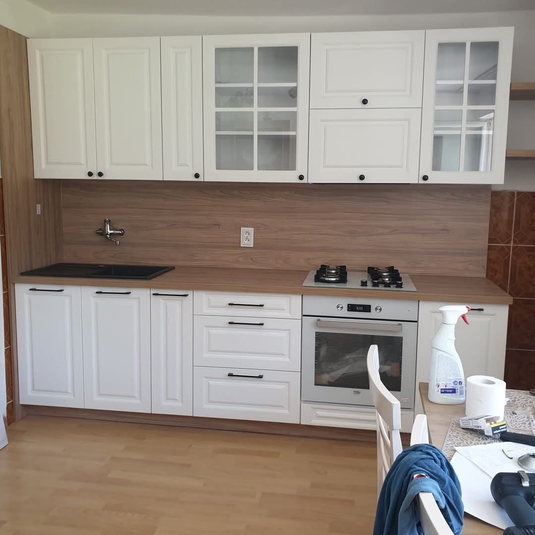 Biała kuchnia z drewnianym blatem i płyta grzewczą. Górne szafki częściowo przeszklone. Widoczny fragment stołu z akcesoriami czyszczącymi.