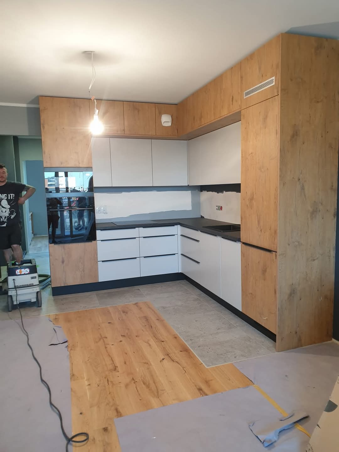 Jasna kuchnia z drewnianymi i białymi frontami, czarnym blatem. Na podłodze panele w trakcie montażu. W tle widoczny odkurzacz przemysłowy i fragment osoby.