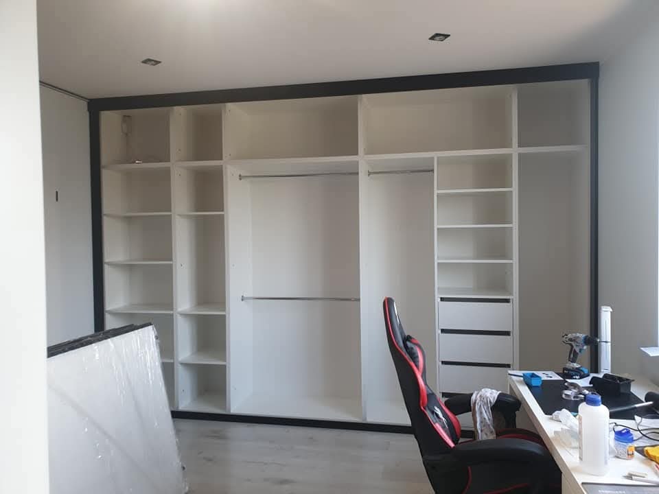 Biała szafa wnękowa z czarną ramą w pokoju w Kalwarii Zebrzydowskiej. Widoczne półki, drążki i biurko z krzesłem gamingowym. Częściowo zapakowane elementy szafy.