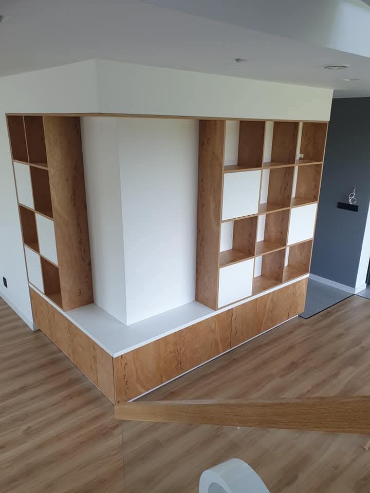 Wnękowa zabudowa z półkami i szafkami z forniru o ciepłym odcieniu drewna, wkomponowana w narożnik pomieszczenia. Białe akcenty na frontach i półkach.