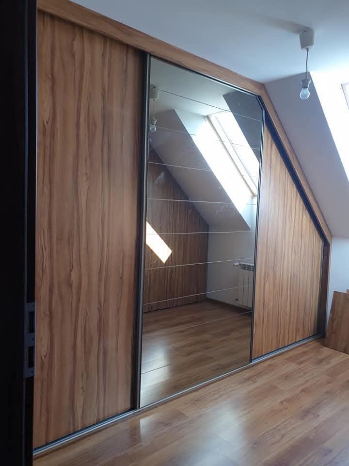 Szafa wnękowa z lustrzanymi drzwiami przesuwnymi, drewnopodobne wykończenie, dopasowana do skosu poddasza, widoczne odbicie okna dachowego.