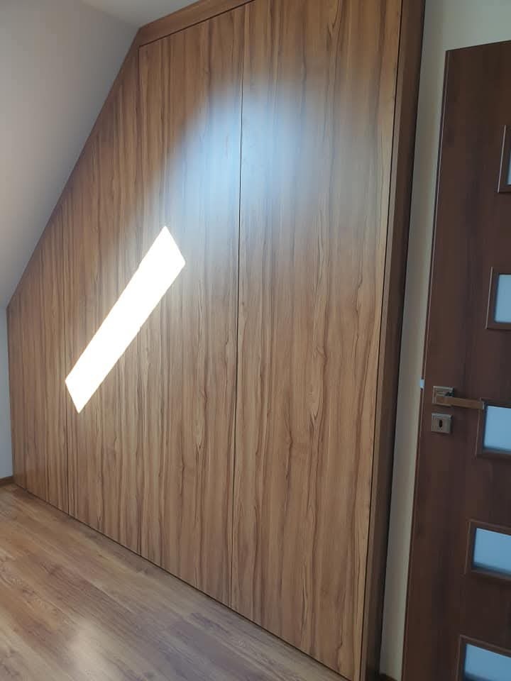 Drewniana szafa wnękowa na wymiar, dopasowana do skosu poddasza. Widoczny fragment drzwi po prawej stronie. Naturalne światło podkreśla fakturę drewna.