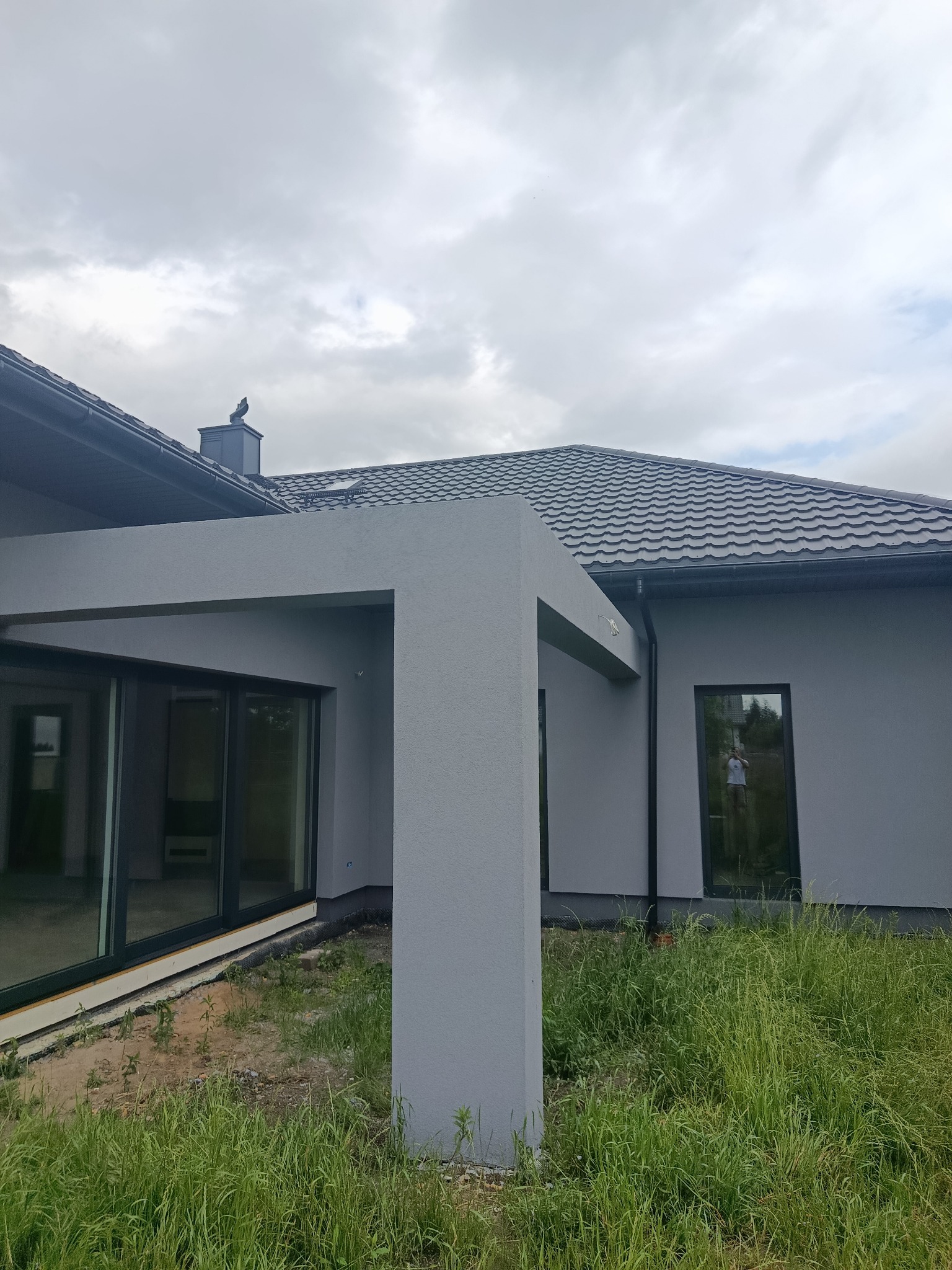 Szara elewacja domu jednorodzinnego z nowoczesną bryłą, widok z zewnątrz, zarośnięty trawnik, okna z czarnymi ramami, pochmurne niebo.