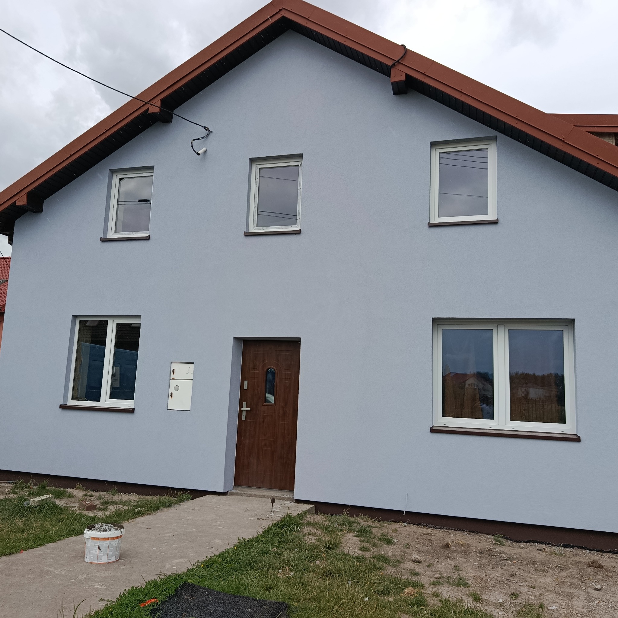 Pomalowana na szaro elewacja domu jednorodzinnego z brązowym dachem. Widoczny fragment chodnika i wiadro po farbie. Białe ramy okien i brązowe drzwi wejściowe.
