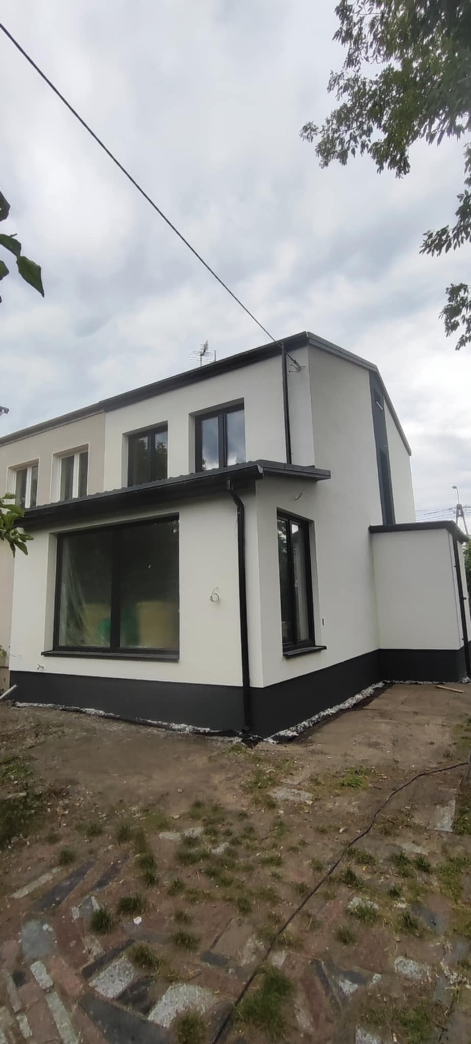 Dwukondygnacyjny dom po renowacji elewacji, z białymi ścianami i czarnymi akcentami. Widoczne okna, rynny i fragment terenu wokół budynku. Ujęcie z dołu.