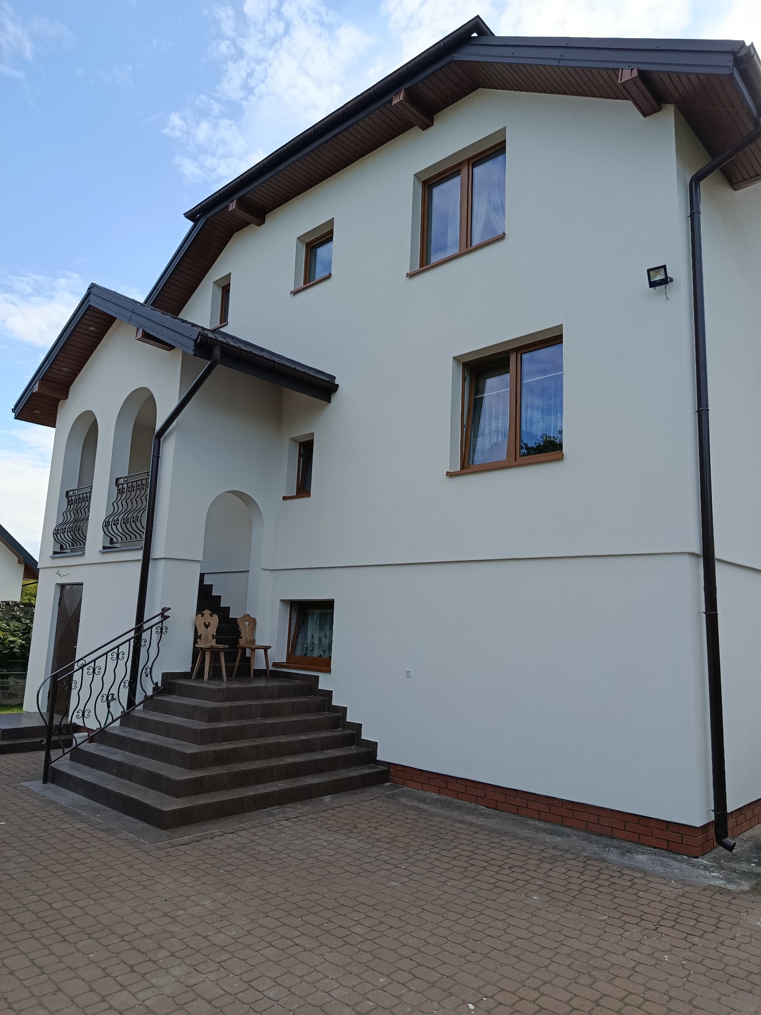 Biała elewacja domu z brązowymi oknami i dachem, widoczne schody wejściowe z balustradą i dekoracyjnymi krzesłami. Jasne niebo w tle.