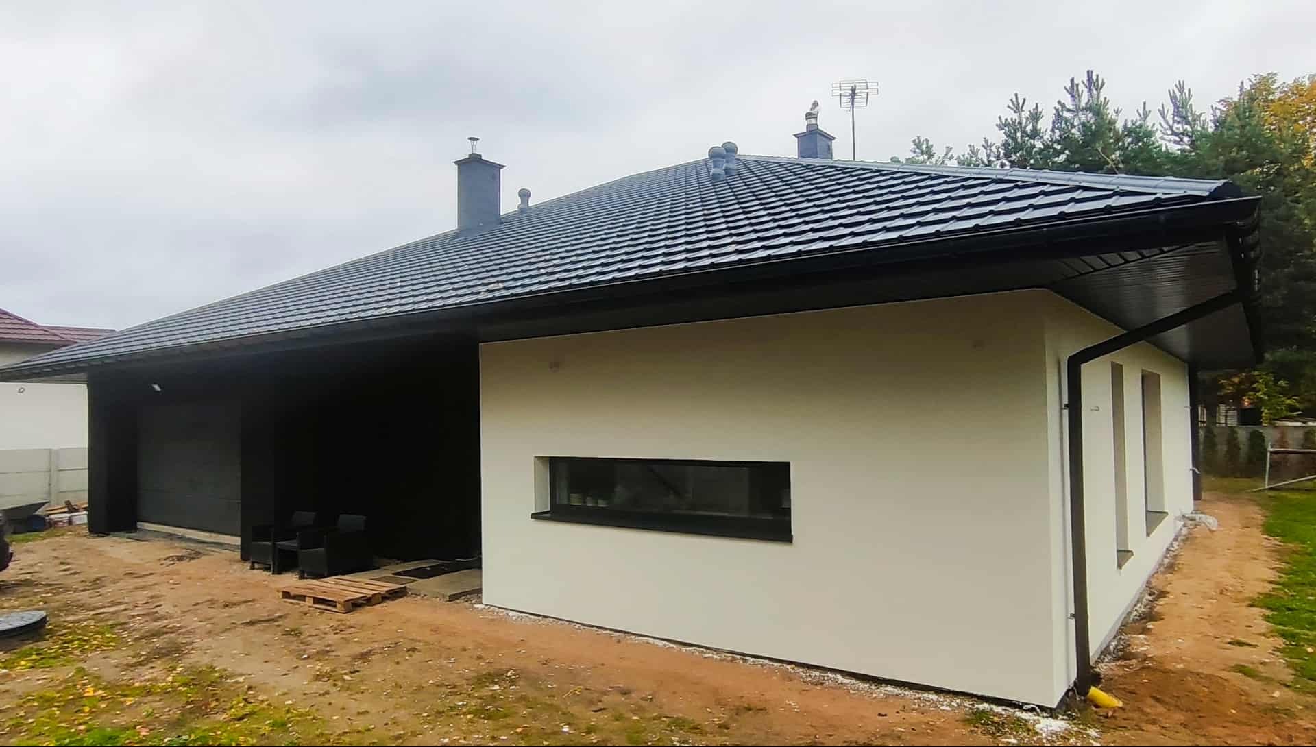Nowoczesny dom jednorodzinny z czarnym dachem i jasną elewacją, widok z boku. Minimalistyczny design, małe okno, czarne rynny. Otoczenie w trakcie prac wykończeniowych.