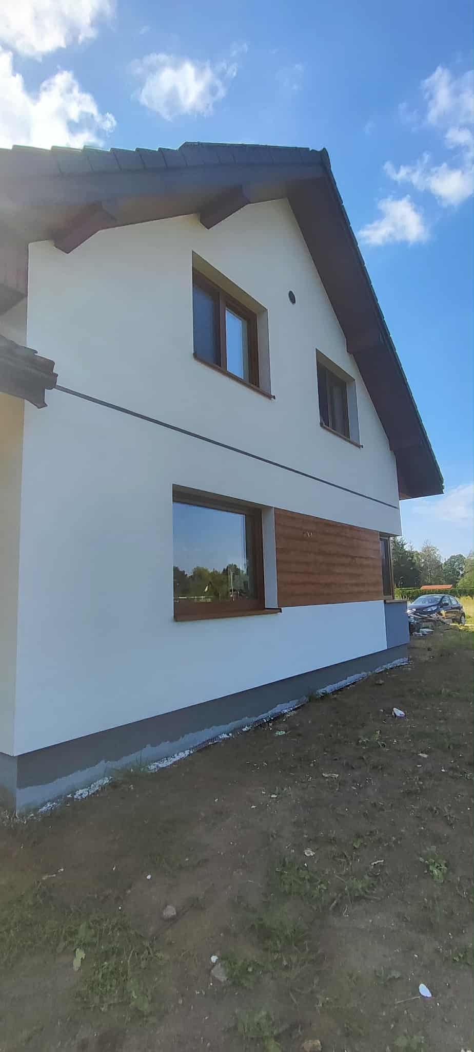 Boczny widok domu po malowaniu elewacji na biało z brązowymi oknami i drewnianą wstawką. Widoczne świeżo pomalowane fundamenty i fragment ogrodu.