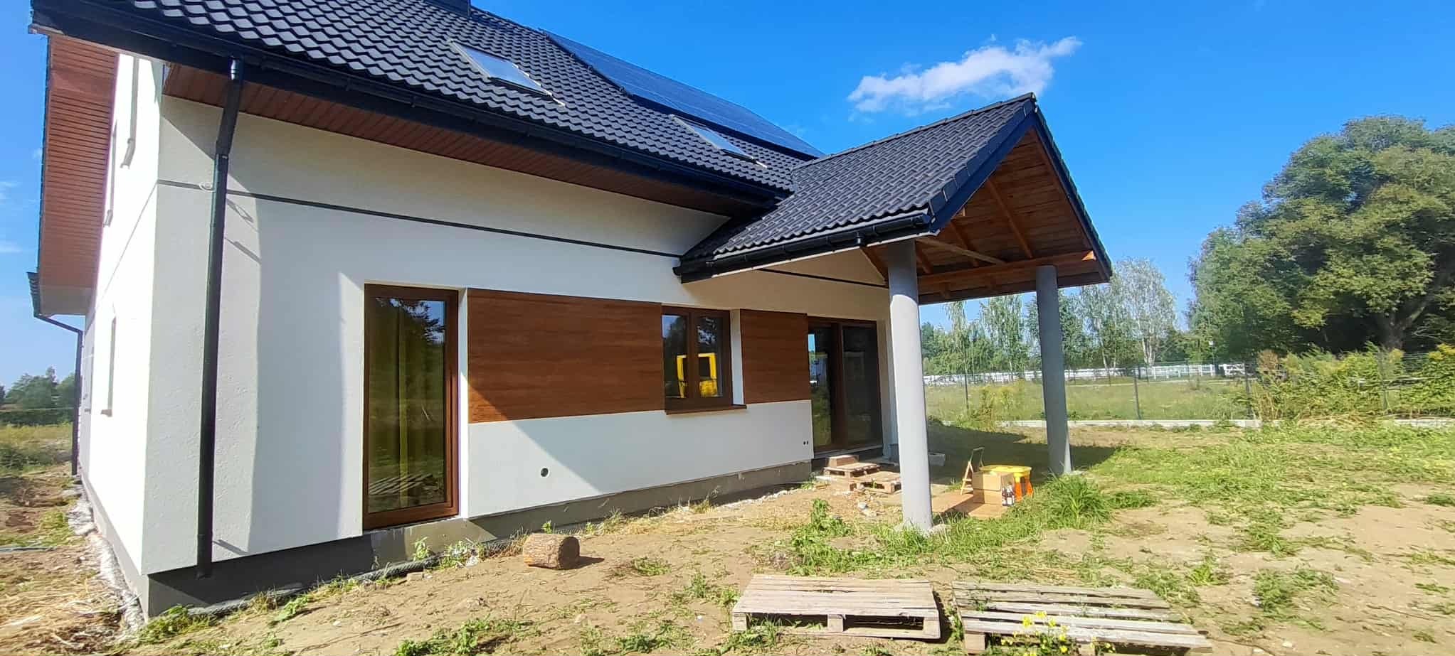 Biała elewacja domu jednorodzinnego z drewnianymi akcentami, czarną dachówką i panelami słonecznymi. Widoczne okna, podcień z kolumnami i teren wokół budynku.