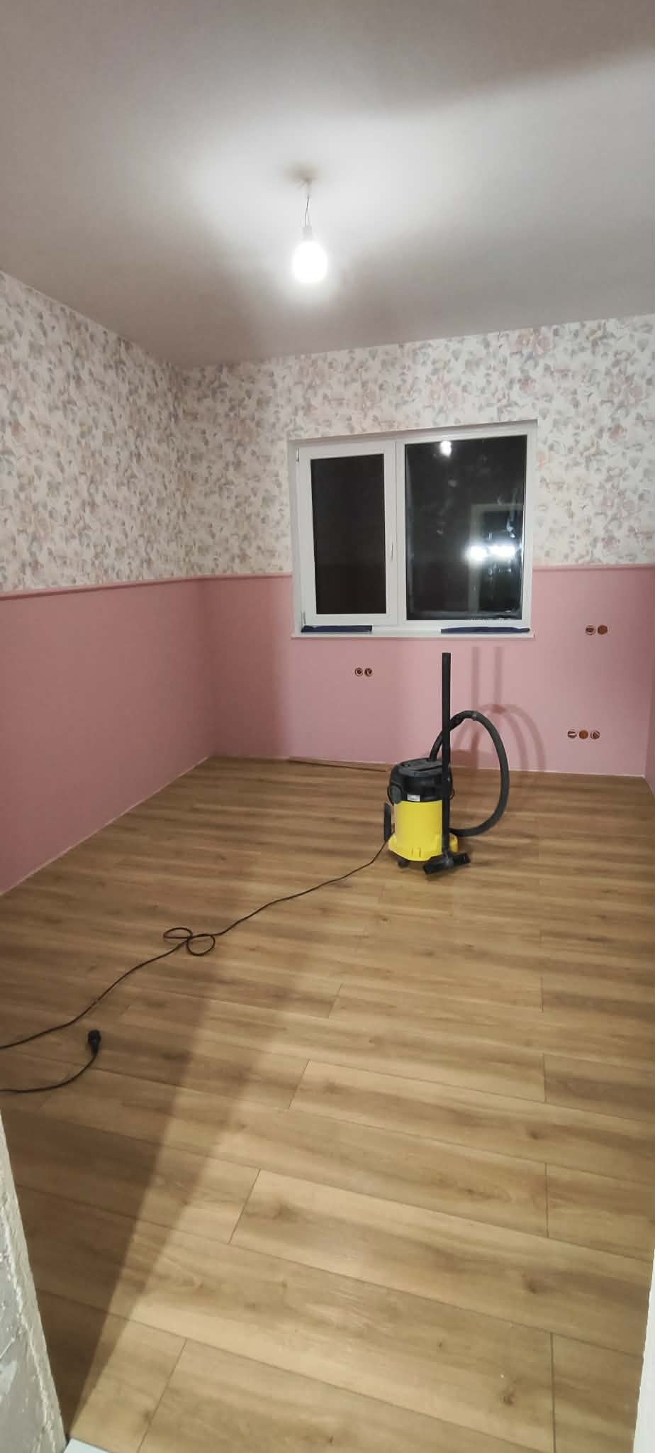 Pokój po remoncie: panele podłogowe, pomalowane ściany do połowy na różowo, tapeta w kwiaty. Żółty odkurzacz na środku. Widok na okno.