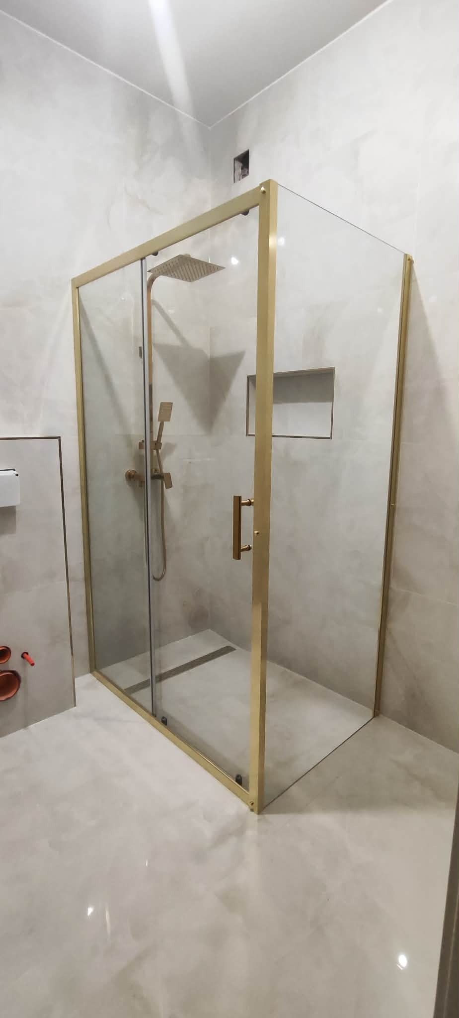 Nowoczesna kabina prysznicowa z złotymi profilami i deszczownicą, w jasnej łazience z marmurowymi płytkami. Widoczna wnęka w ścianie.