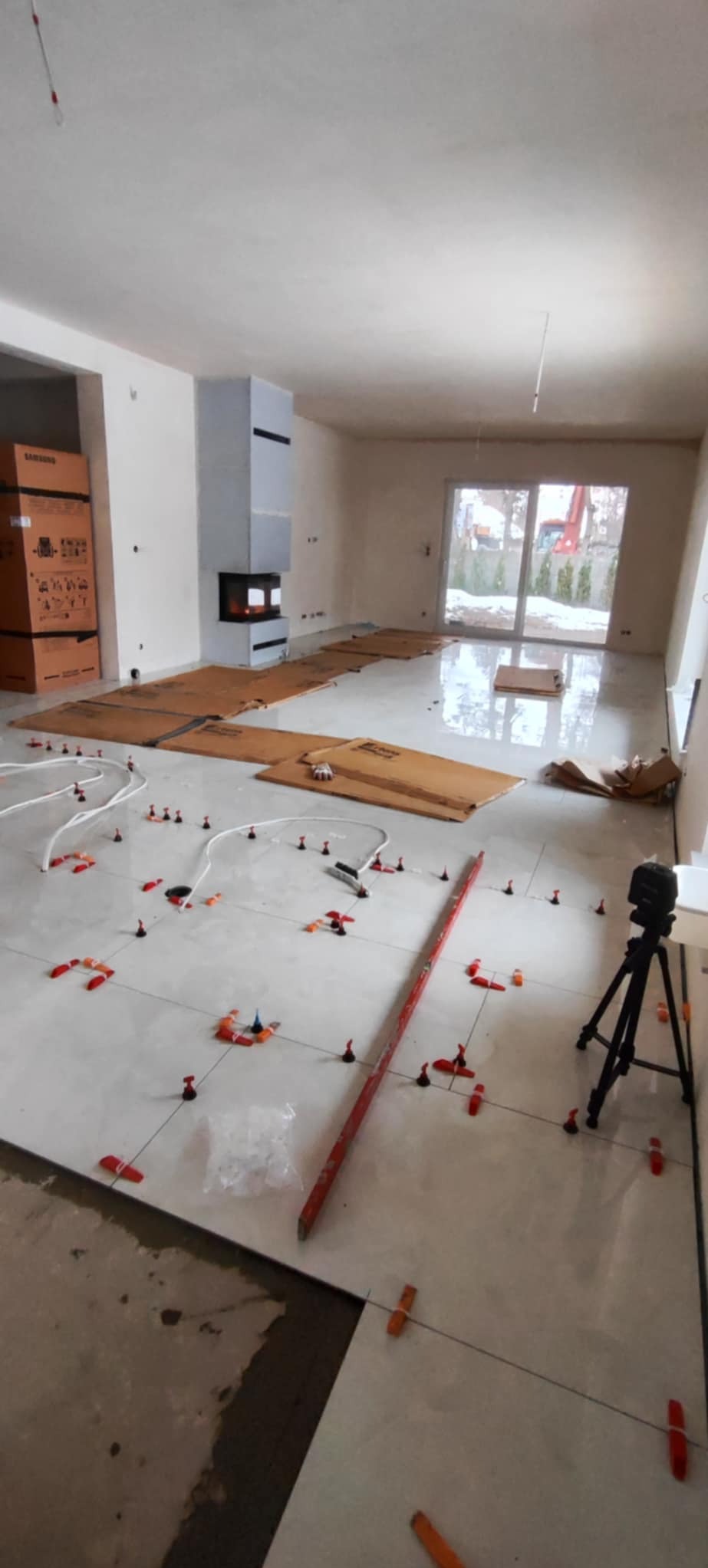 Układanie jasnych płytek podłogowych w salonie z kominkiem. Widoczne narzędzia: poziomica, laser krzyżowy, system poziomowania płytek. Częściowo zabezpieczona podłoga kartonem.