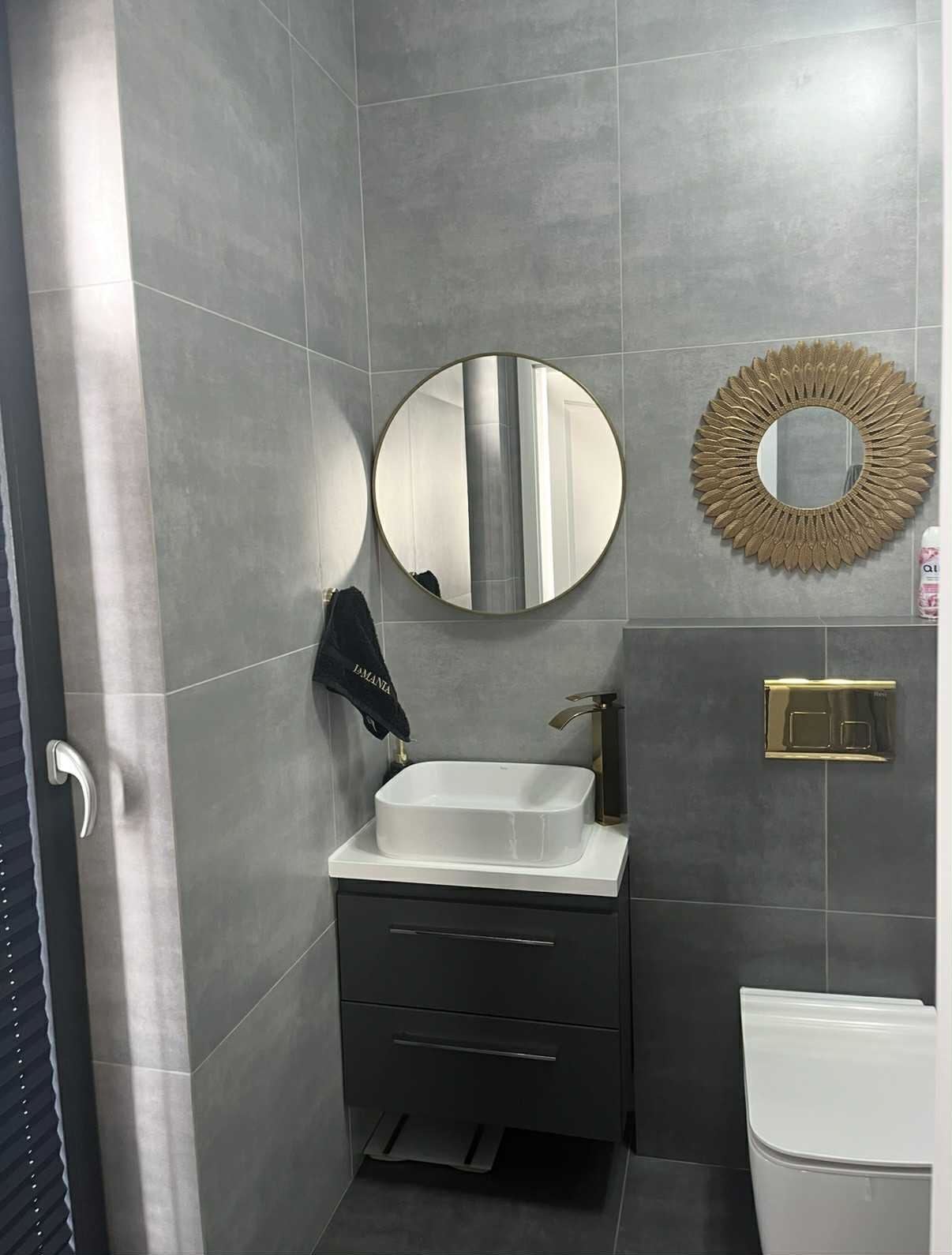 Nowoczesna łazienka z szarymi płytkami, okrągłymi lustrami i białą umywalką na czarnej szafce. Złota bateria i przycisk spłuczki dodają elegancji. Minimalistyczny design.