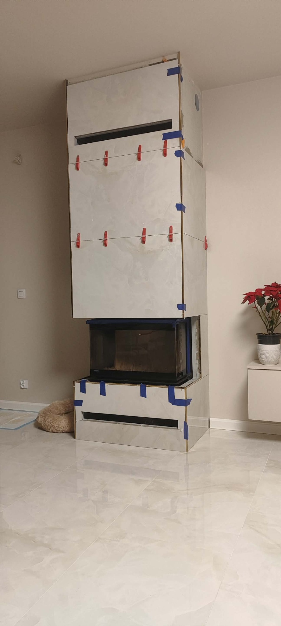 Obudowa kominka w trakcie wykańczania, z widocznymi płytkami mocowanymi za pomocą klipsów i taśmy. W tle jasna ściana i podłoga z płytek.