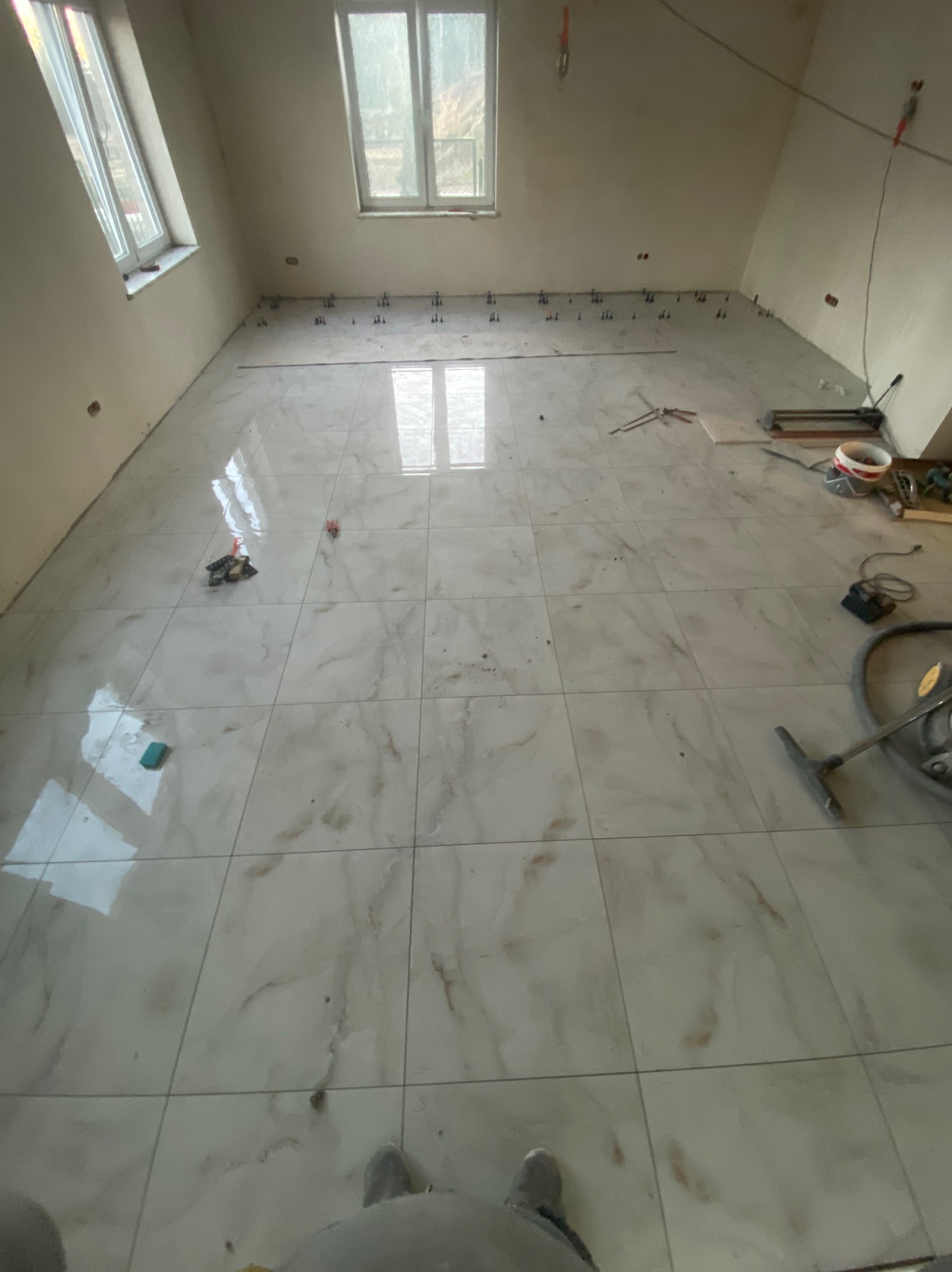 Układanie jasnych, marmurkowych płytek podłogowych w pokoju. Widoczne narzędzia glazurnicze i krzyżyki dystansowe. Dwa okna w skośnej ścianie.