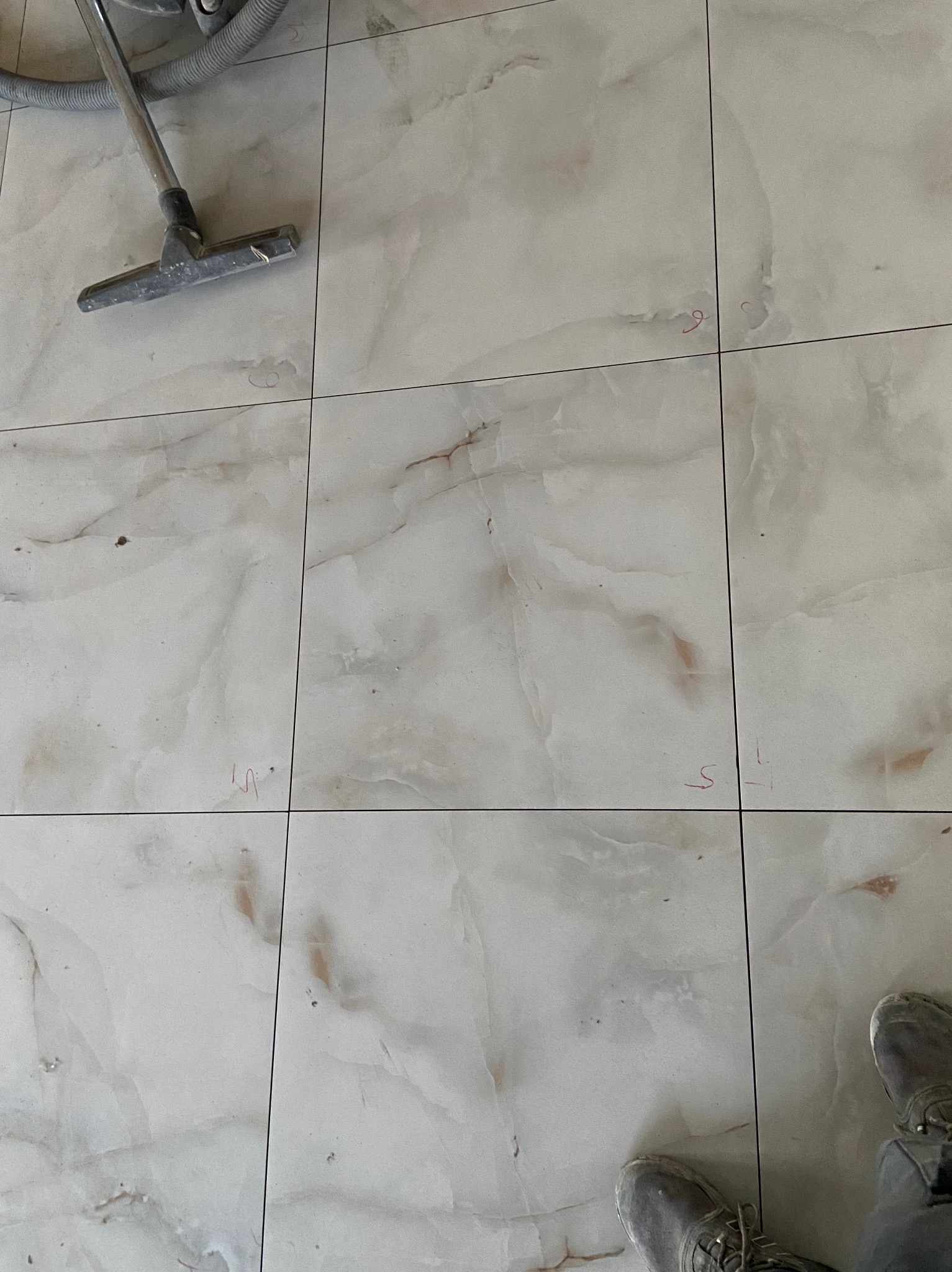 Jasne, marmurowe płytki podłogowe po ułożeniu, z widocznymi śladami po czyszczeniu i oznaczeniami. W rogu widoczny fragment odkurzacza i butów.
