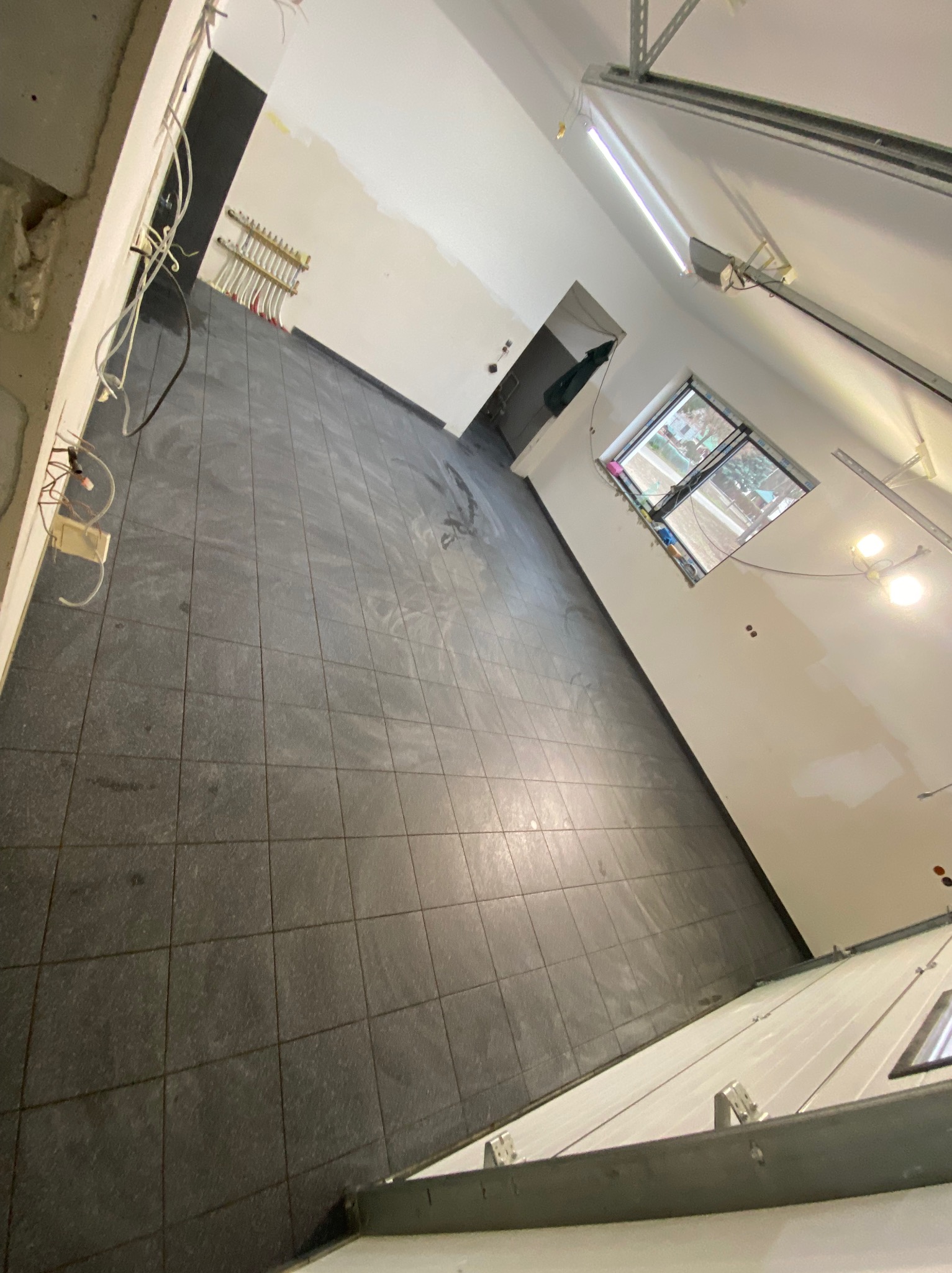 Ułożona, ciemnoszara podłoga z płytek w nowym pomieszczeniu. Widoczne kable, świeżo pomalowane ściany i okno z widokiem na zewnątrz. Całość w surowym stanie.