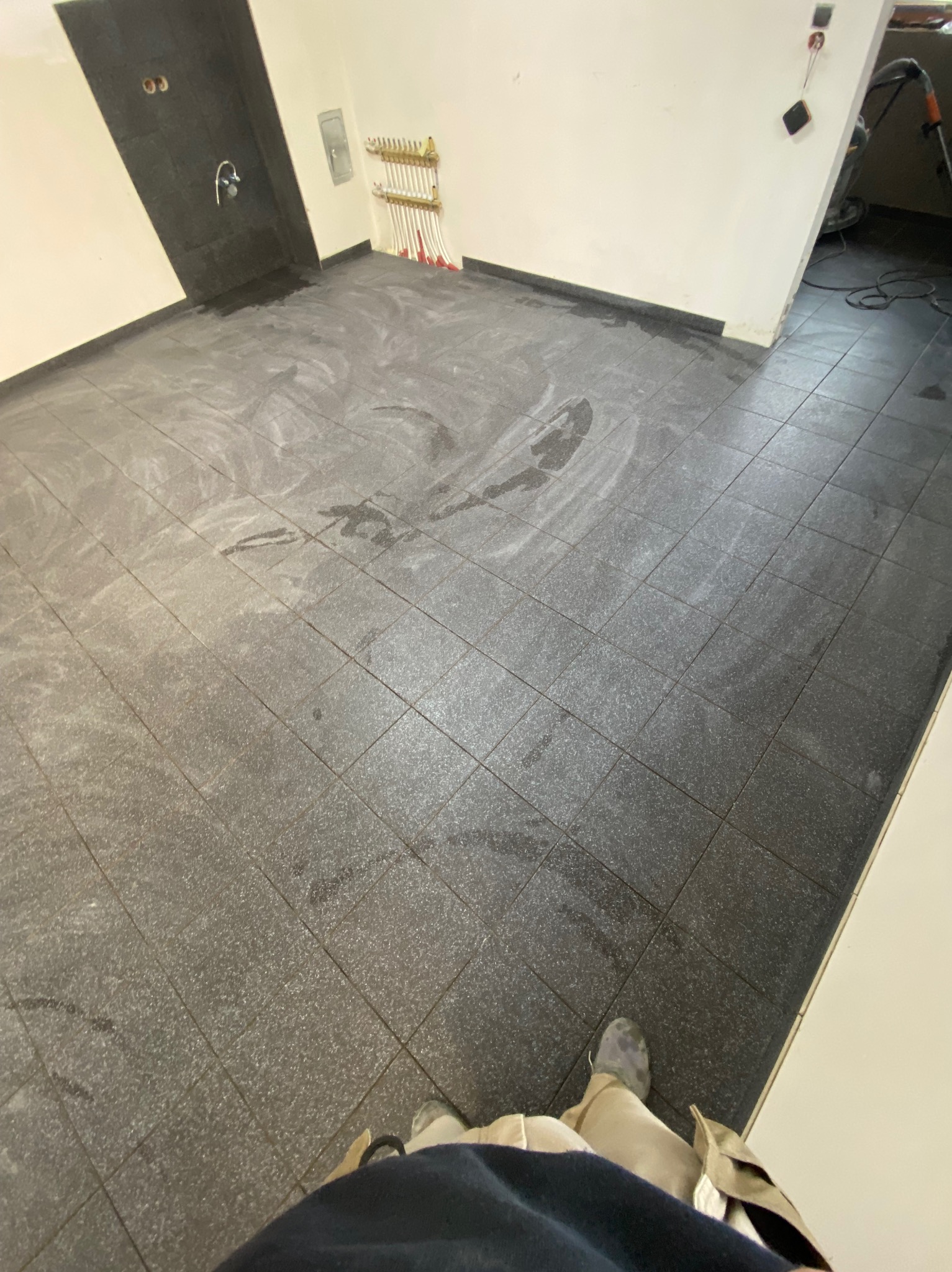 Ułożona, ciemnoszara podłoga z płytek w łazience, widoczne ślady po fugowaniu. Grzejnik drabinkowy na ścianie, fragment osoby w kadrze.