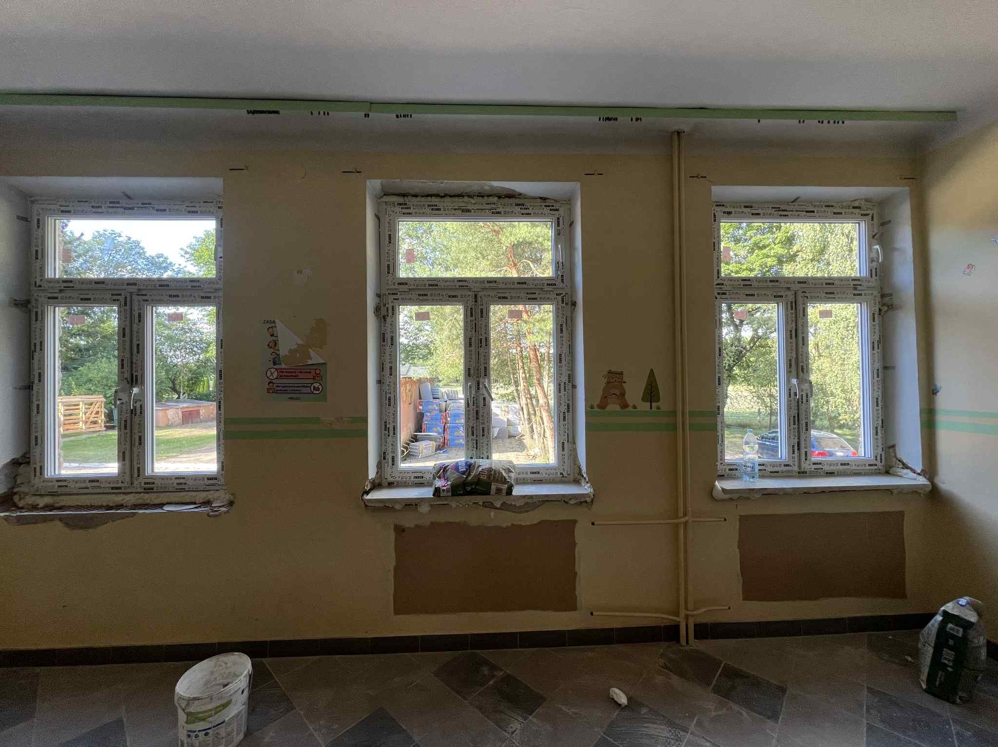 Trzy okna PCV w trakcie montażu/obróbki w pomieszczeniu z szarą podłogą. Na ścianie widoczne ślady po pracach wykończeniowych, wiadro z farbą i worek z zaprawą.