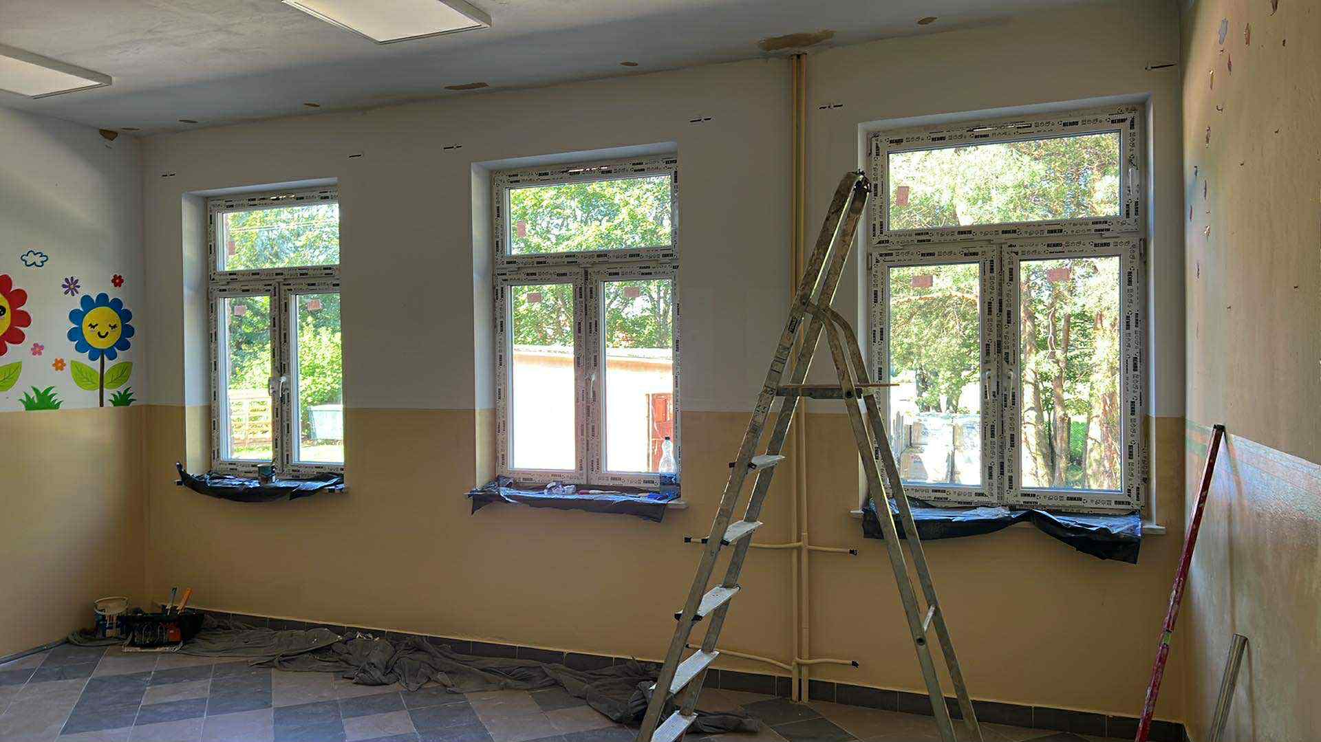 Pokój po malowaniu z naklejkami kwiatów, drabina oparta o ścianę, zabezpieczone okna folią, widoczne ślady farby na ścianie i podłodze, w tle zieleń za oknem.