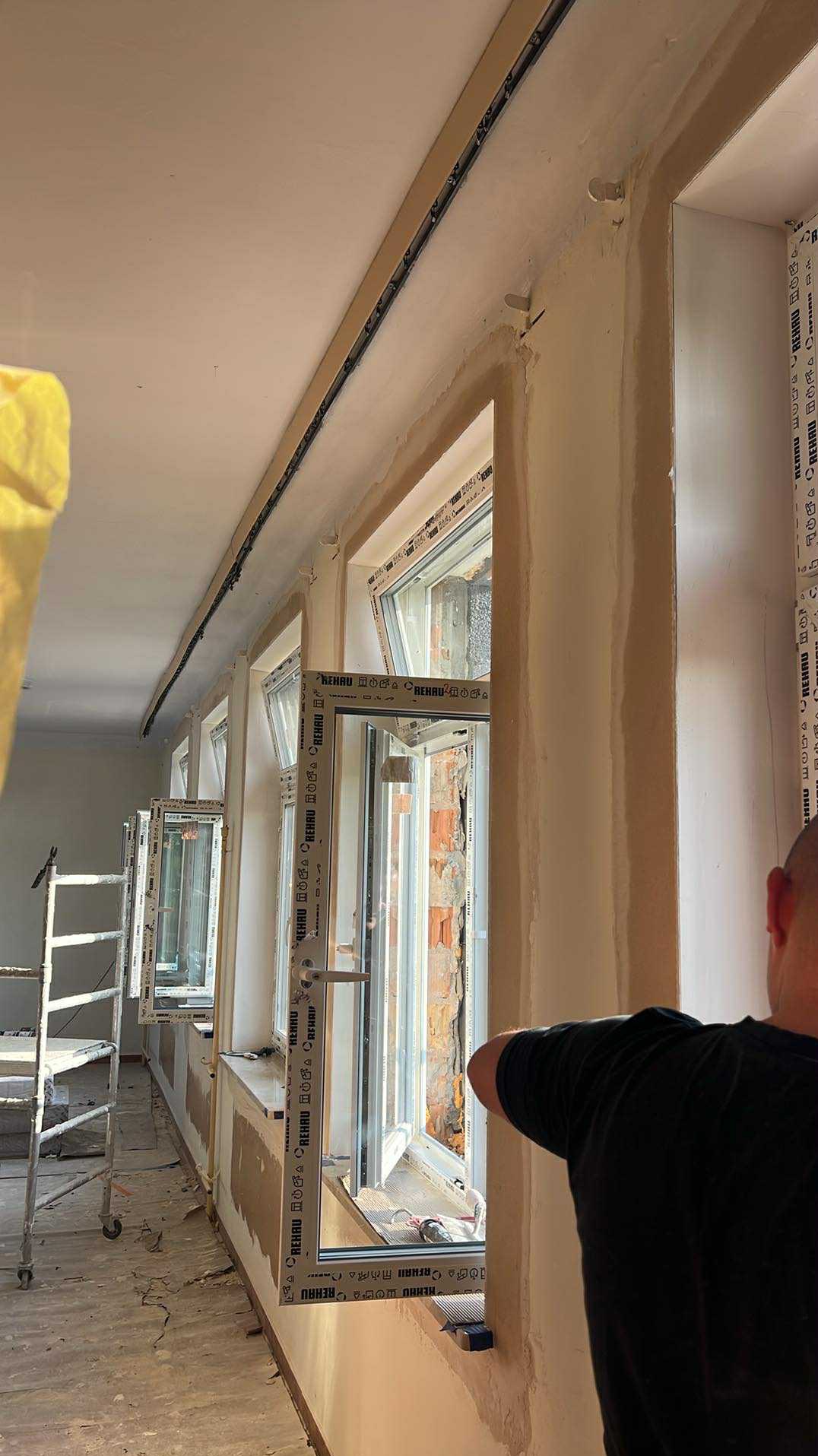 Montaż okien PCV Rehau w nowym budynku, widoczne przygotowane ściany do szpachlowania i drabina. Częściowo widoczna osoba w czarnej koszulce.