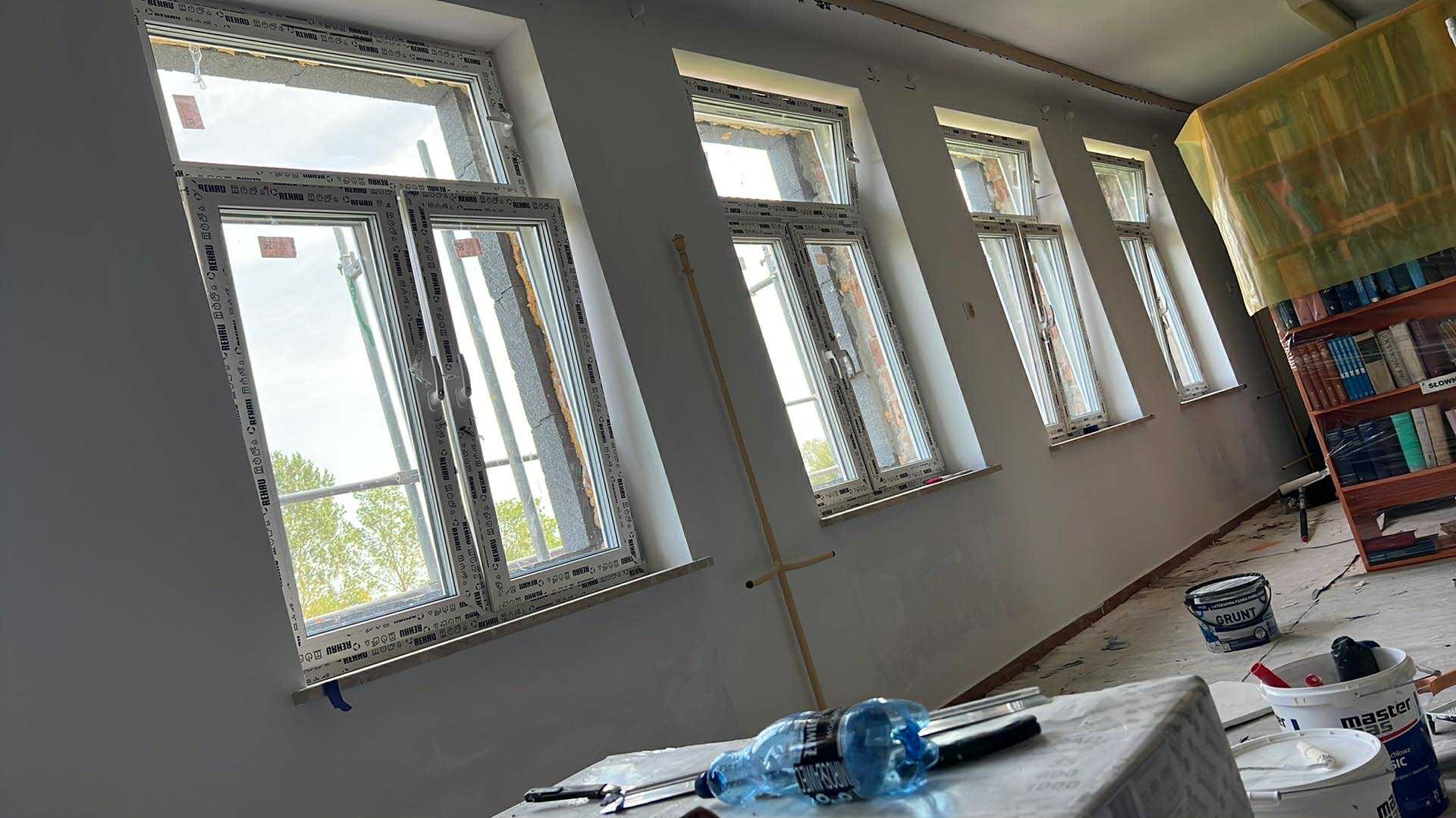 Wnętrze w trakcie remontu: rząd okien z białymi ramami, częściowo osłonięty regał z książkami, wiaderka z farbą i narzędzia malarskie na pierwszym planie.