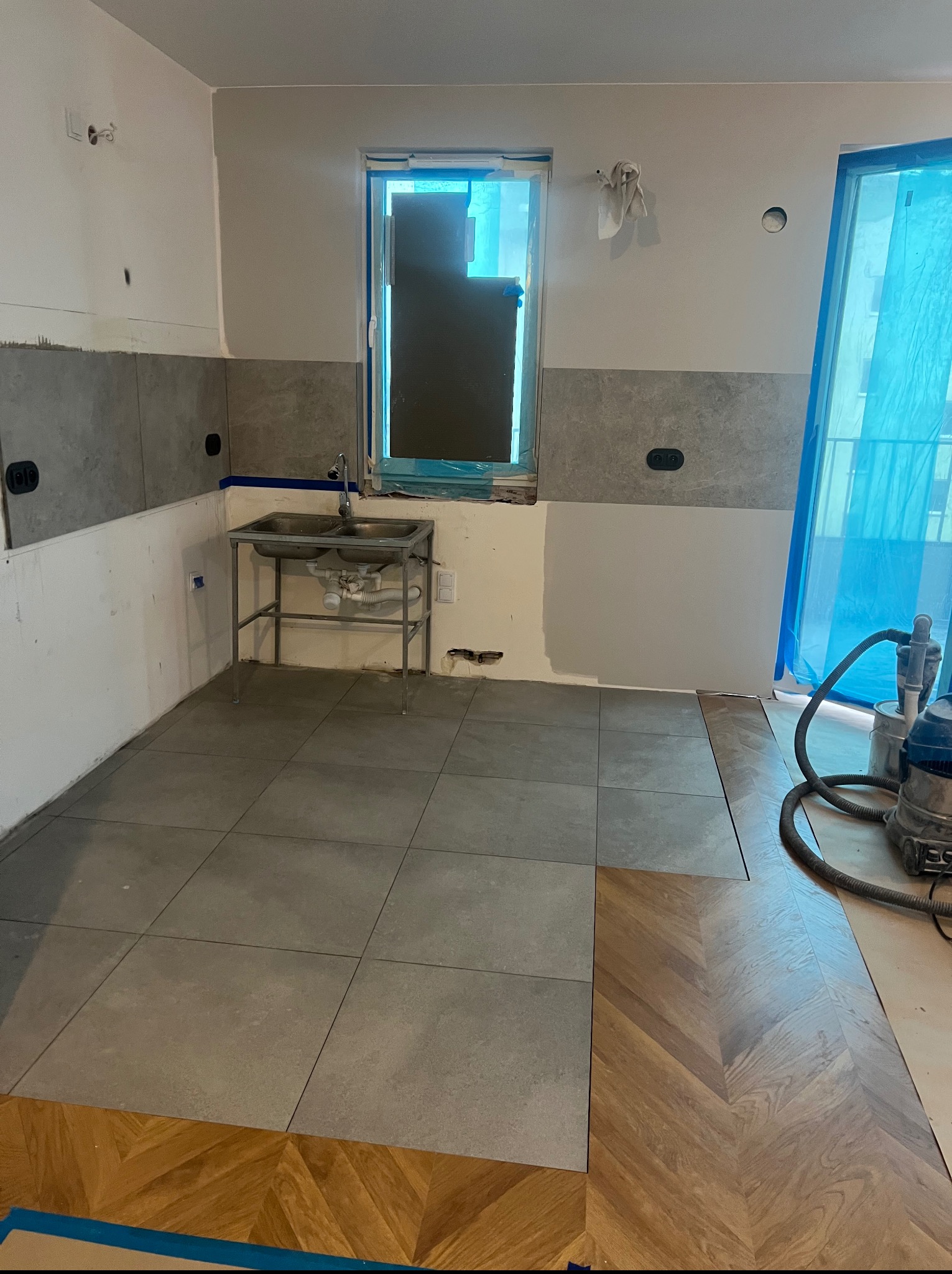 Wnętrze kuchni w trakcie remontu: częściowo ułożone płytki podłogowe i parkiet w jodełkę, zabezpieczone okno, widoczny odkurzacz przemysłowy i metalowy zlew.