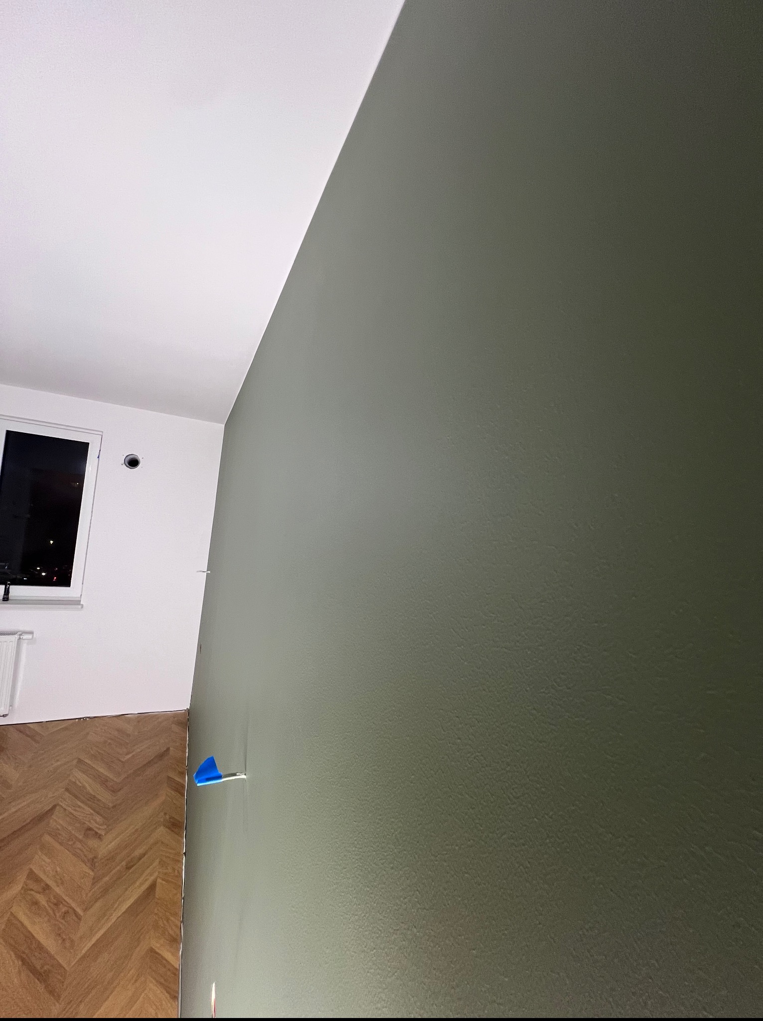 Ściana pomalowana na oliwkowy kolor, widoczny fragment parkietu i okno. Na ścianie niebieska taśma malarska. Ukończone malowanie mieszkania.