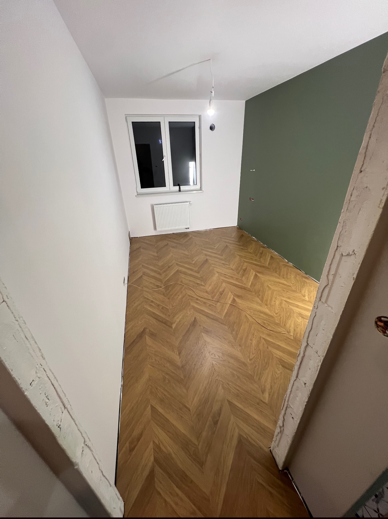 Pokój po remoncie: parkiet w jodełkę, białe ściany i jedna ściana w kolorze butelkowej zieleni. Widoczne okno i grzejnik. Wykończenie w trakcie realizacji.