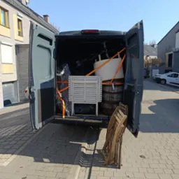 Otwarty bagażnik busa wypełniony meblami i beczką zabezpieczonymi pomarańczowymi pasami transportowymi. W tle zabudowa miejska i błękitne niebo.