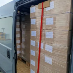 Wnętrze busa wypełnione po brzegi zapakowanymi kartonami, zabezpieczonymi czerwoną taśmą transportową. Widoczna podłoga wyłożona płytami drewnianymi.