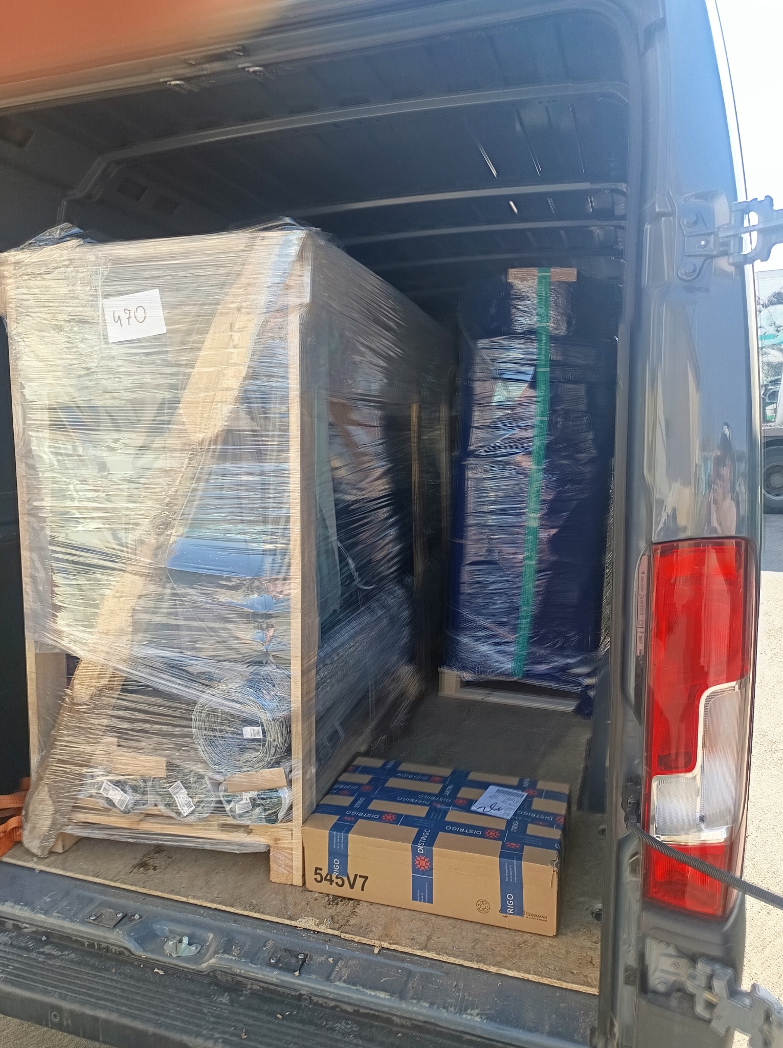 Wnętrze busa z paletami owiniętymi folią stretch, karton z napisem DISTRIGO. Widok od tyłu, otwarte drzwi, światło dzienne. Transport towarów.