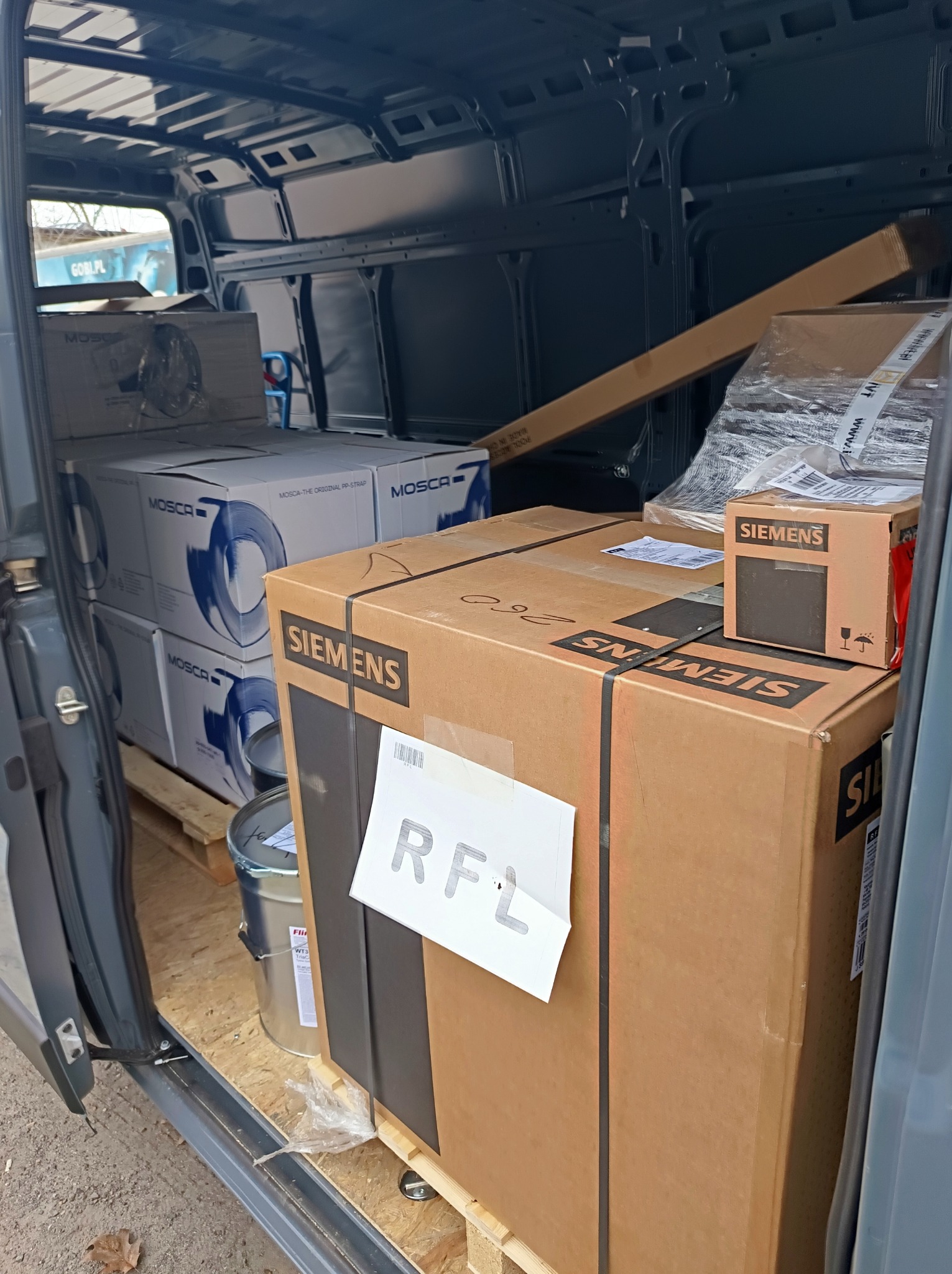 Wnętrze busa wypełnione paletami z kartonami marek Siemens i Mosca, gotowymi do transportu. Widoczne zabezpieczenia ładunku i częściowo otwarte drzwi pojazdu.