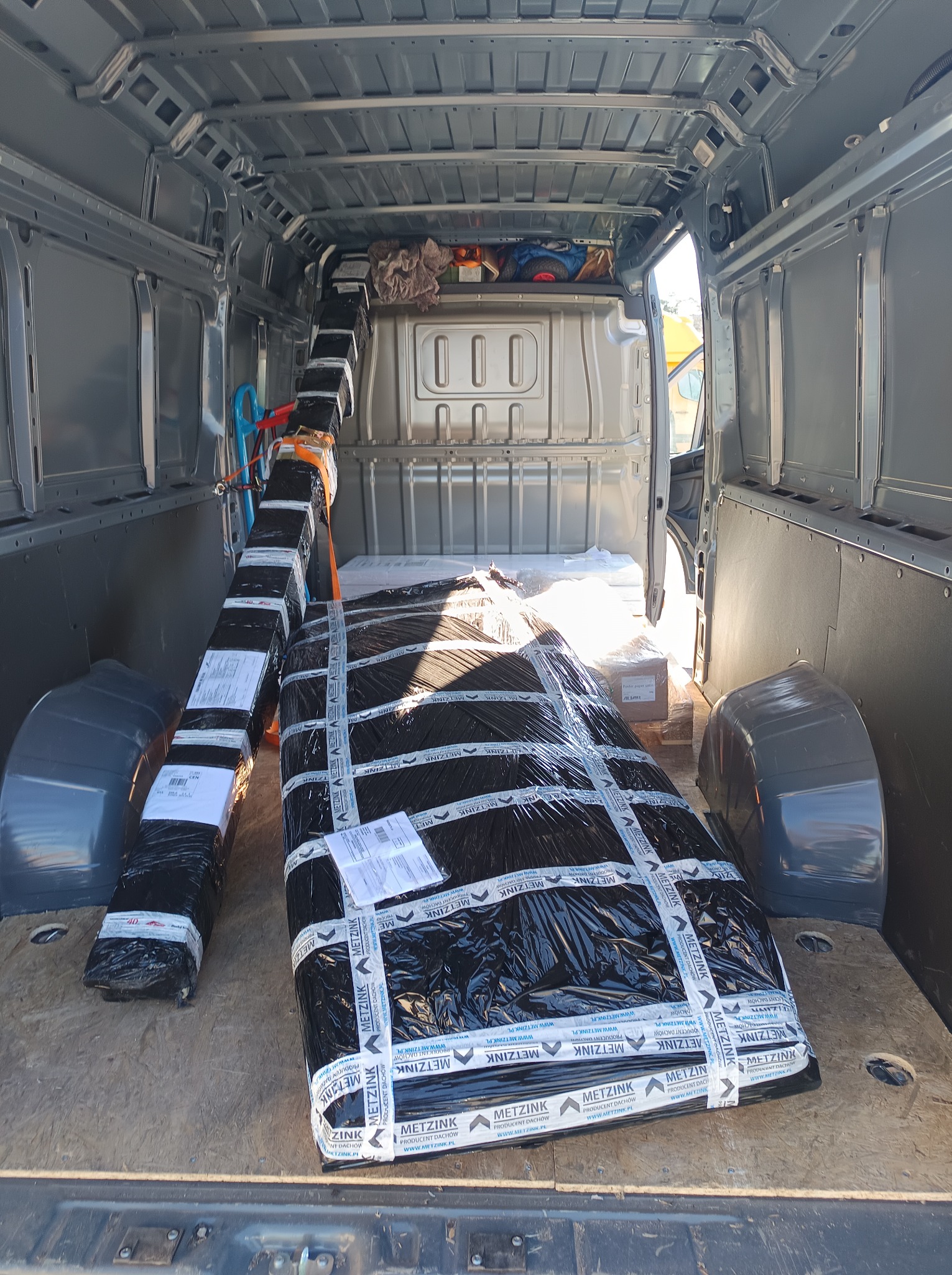 Wnętrze busa wypełnione zapakowanymi elementami, zabezpieczonymi folią i taśmą z logo producenta dachów. Widok z tyłu pojazdu, częściowo widoczne siedzenia kierowcy.