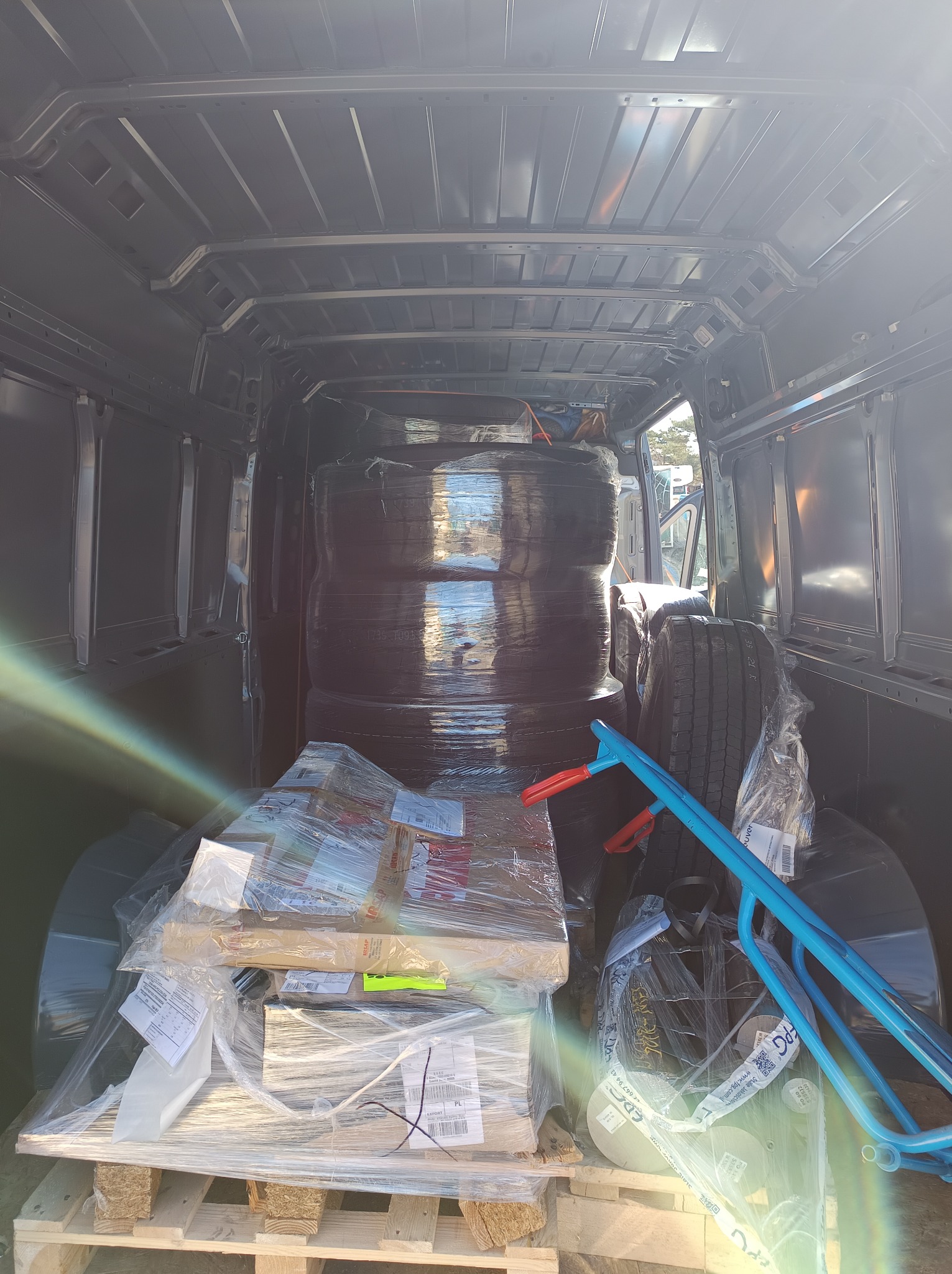 Wnętrze busa wypełnione po brzegi: opony owinięte folią, paczki na palecie, niebieski wózek transportowy. Widok z tyłu pojazdu, jasne światło wpadające z zewnątrz.