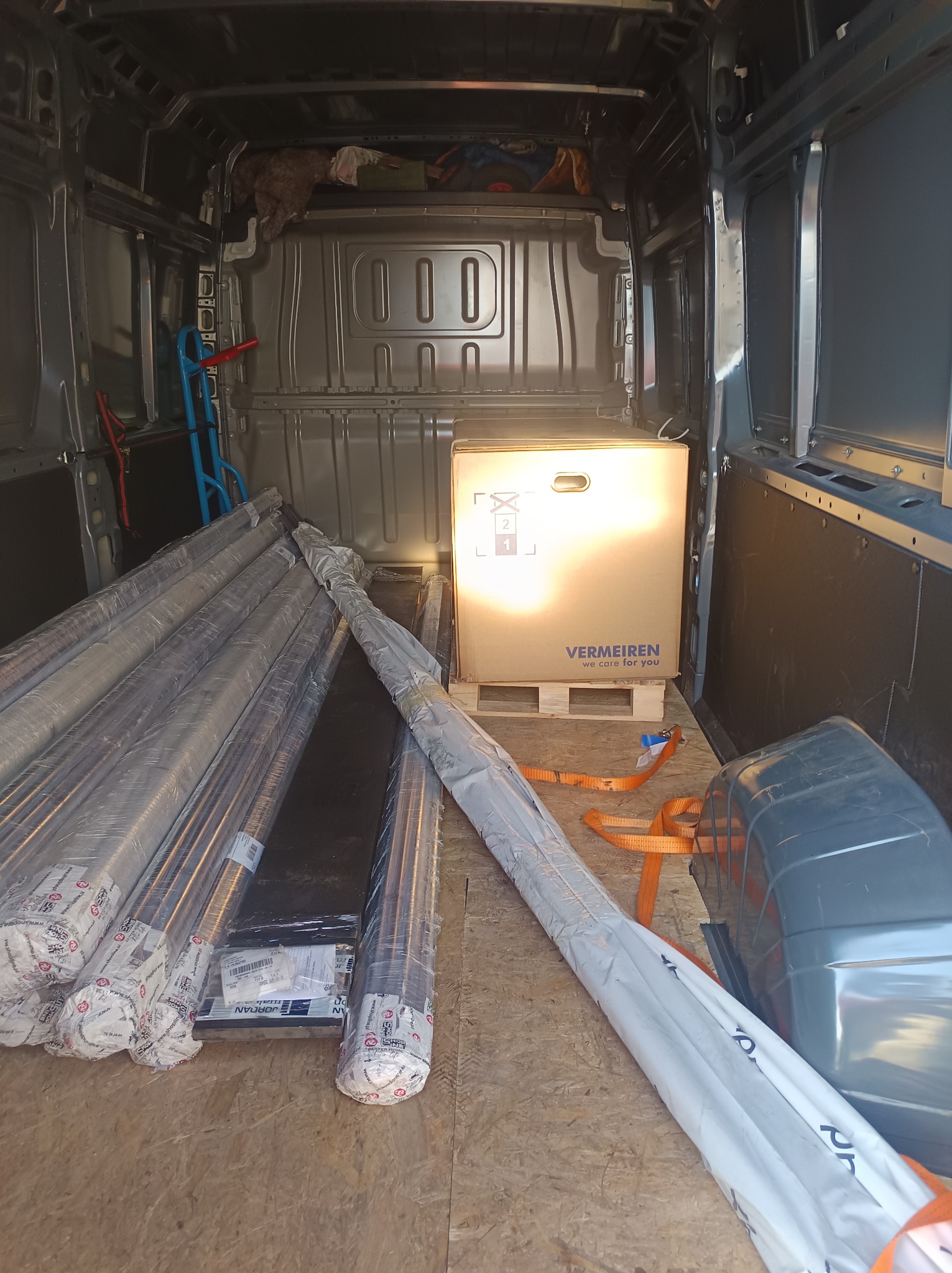 Wnętrze busa z ładunkiem: długie rury w folii, karton na palecie zabezpieczony pasami. Widoczne ścianki pojazdu i podłoga z płyty. Częściowo widoczny wózek transportowy.