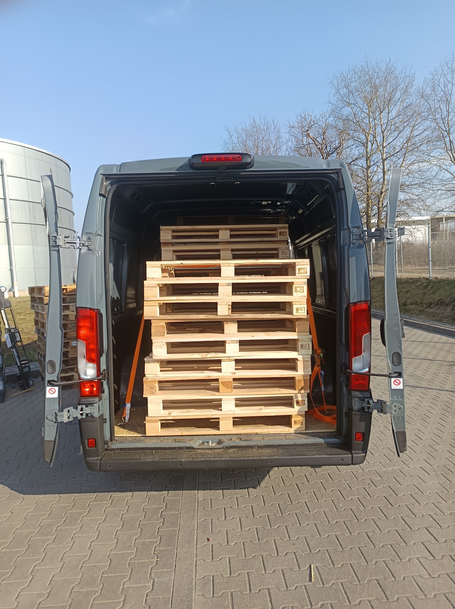 Szary bus z otwartymi drzwiami, załadowany paletami, zabezpieczonymi pomarańczowymi pasami. Widok z tyłu na kostkę brukową i silos w tle.