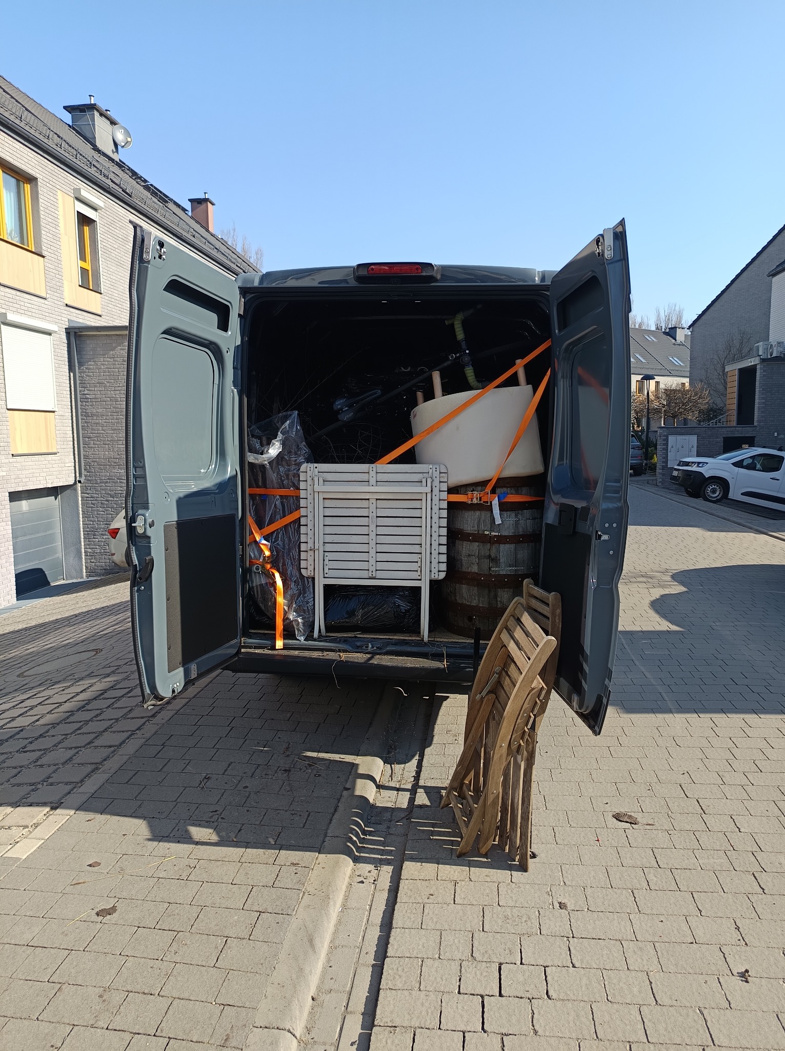 Otwarty bagażnik busa wypełniony meblami i beczką zabezpieczonymi pomarańczowymi pasami transportowymi. W tle zabudowa miejska i błękitne niebo.