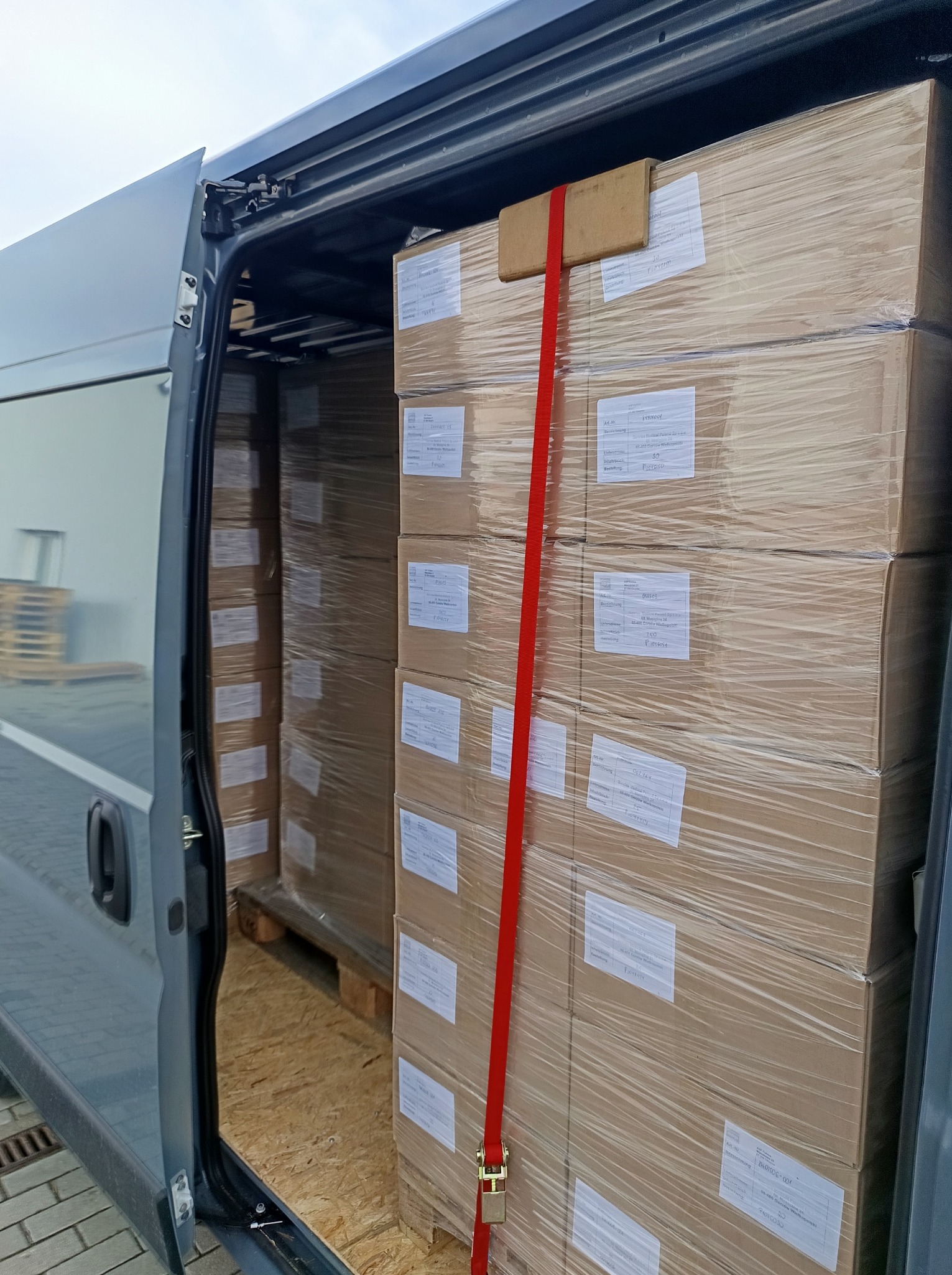 Wnętrze busa wypełnione po brzegi zapakowanymi kartonami, zabezpieczonymi czerwoną taśmą transportową. Widoczna podłoga wyłożona płytami drewnianymi.