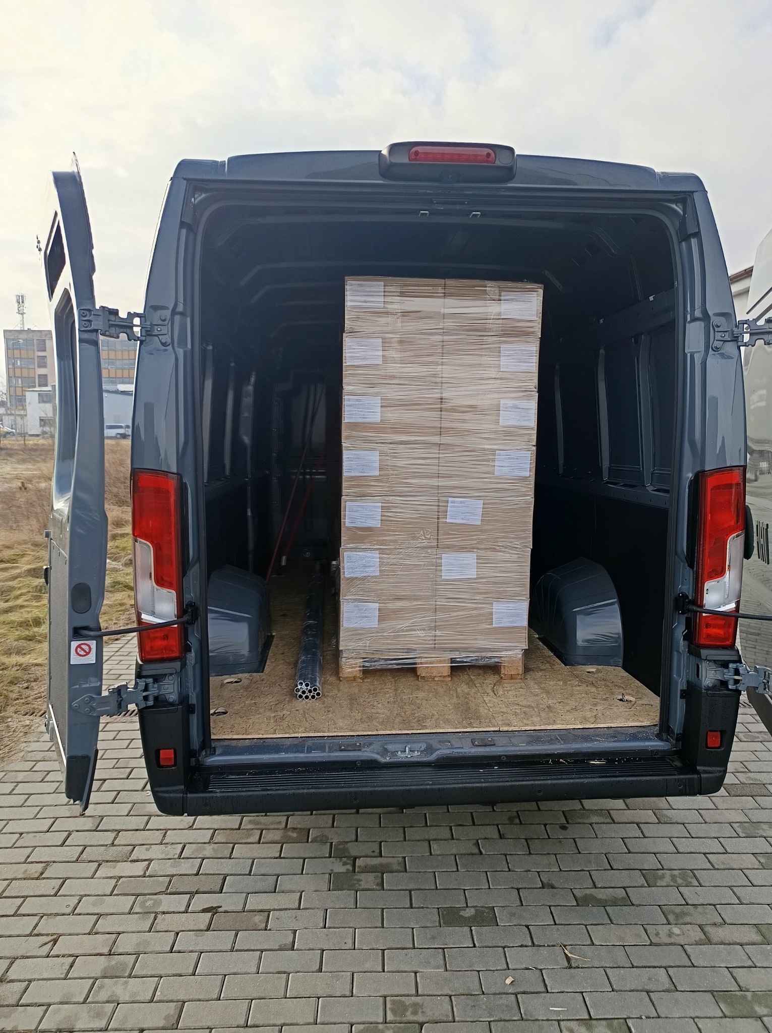 Szary bus z otwartymi drzwiami bagażnika, wewnątrz paleta z zapakowanym towarem. Widoczne rury i elementy wyposażenia. Tło: budynek i kostka brukowa.