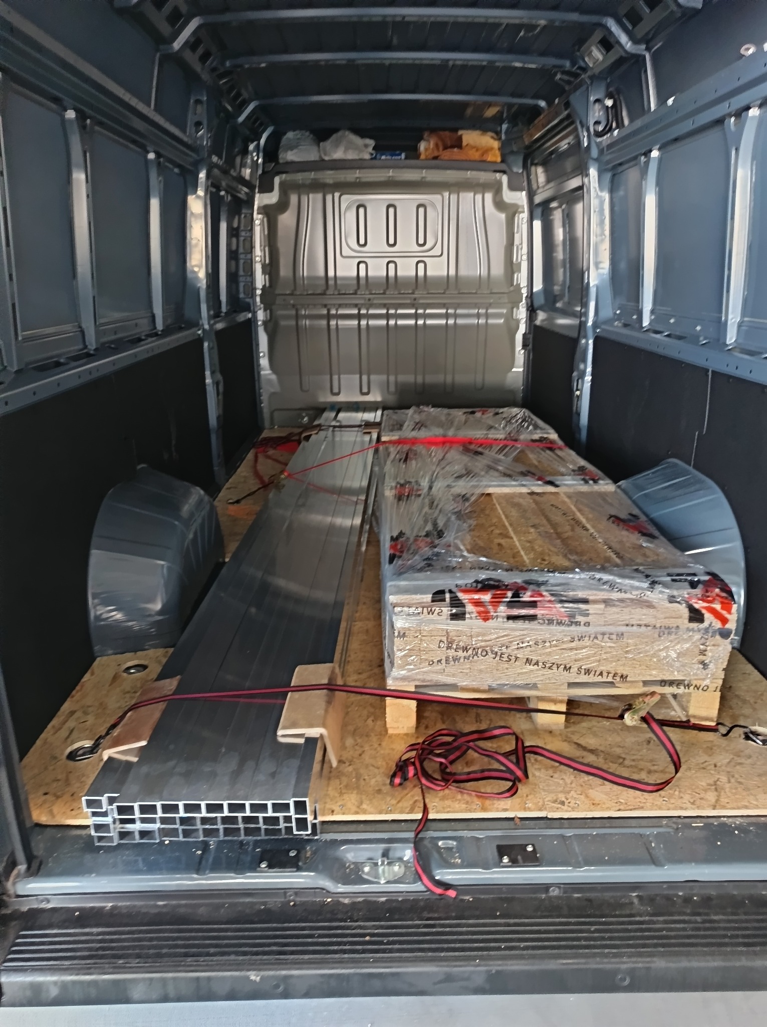 Wnętrze busa z ładunkiem: profile aluminiowe i paleta drewna zabezpieczone pasami transportowymi. Widok z tyłu pojazdu, oświetlenie dzienne.