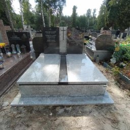 Kamil Jaszczyński - Podwójny nagrobek z granitu Viscount White i Premium Black na cmentarzu, z inskrypcjami i krzyżem, otoczony zielenią i innymi nagrobkami. Błyszcząca powierzchnia odbija niebo.