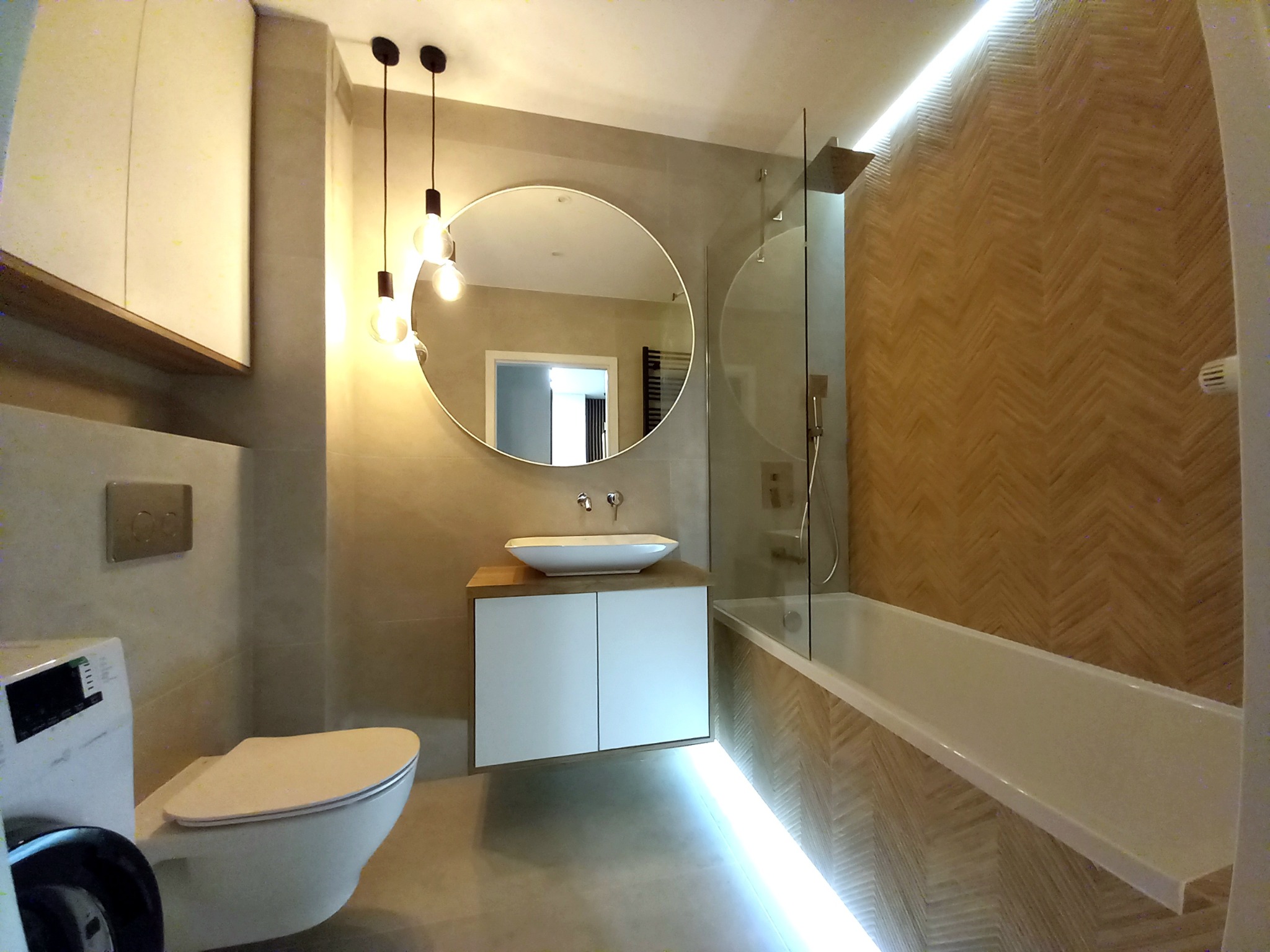 Nowoczesna łazienka z okrągłym lustrem, wiszącą szafką z umywalką i podświetlaną wanną, wykończona płytkami imitującymi drewno ułożonymi w jodełkę.