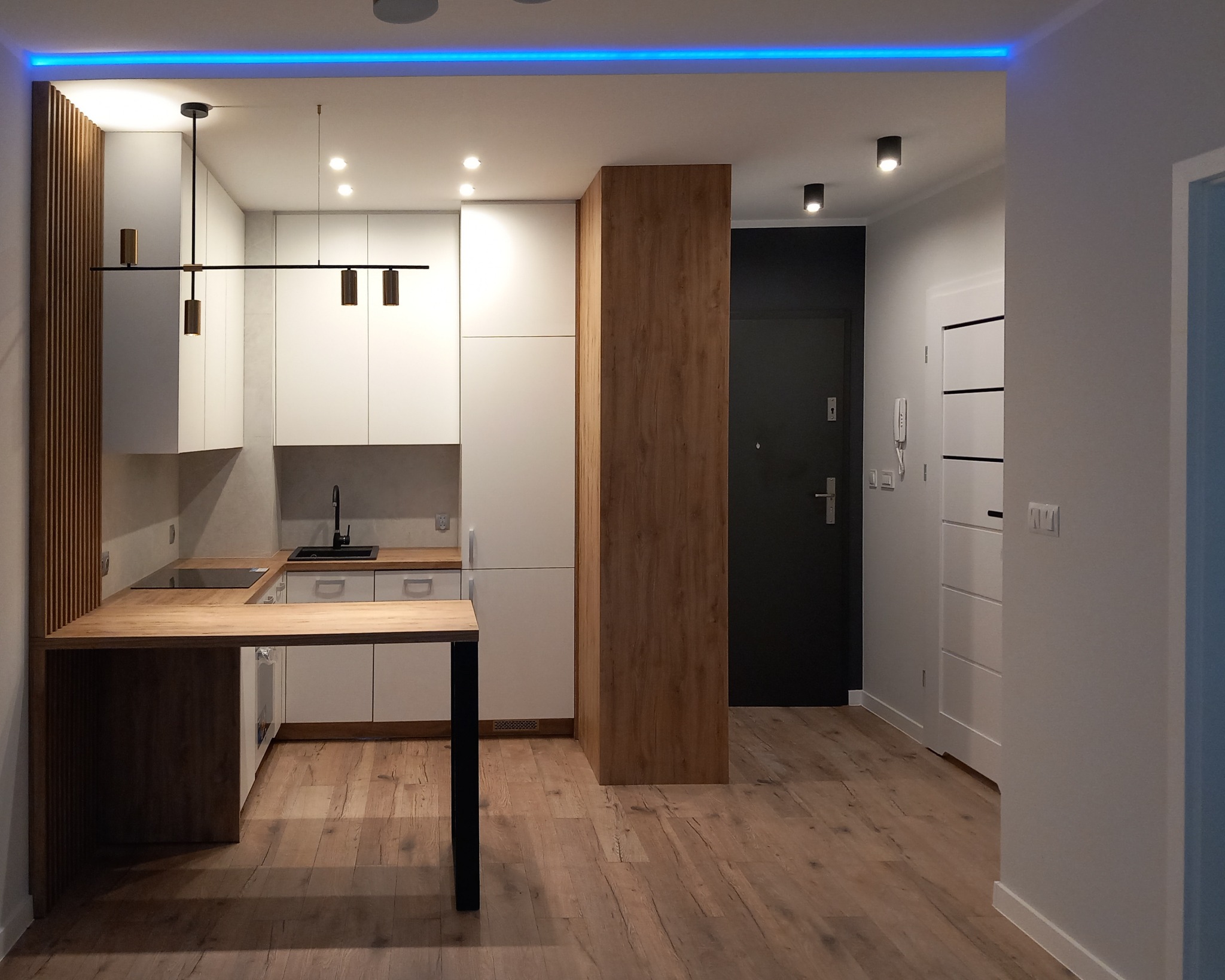 Nowoczesna kuchnia z białymi szafkami, drewnianym blatem i podświetleniem LED. Widok na przedpokój z czarnymi drzwiami i białymi szafami. Parkiet na podłodze.