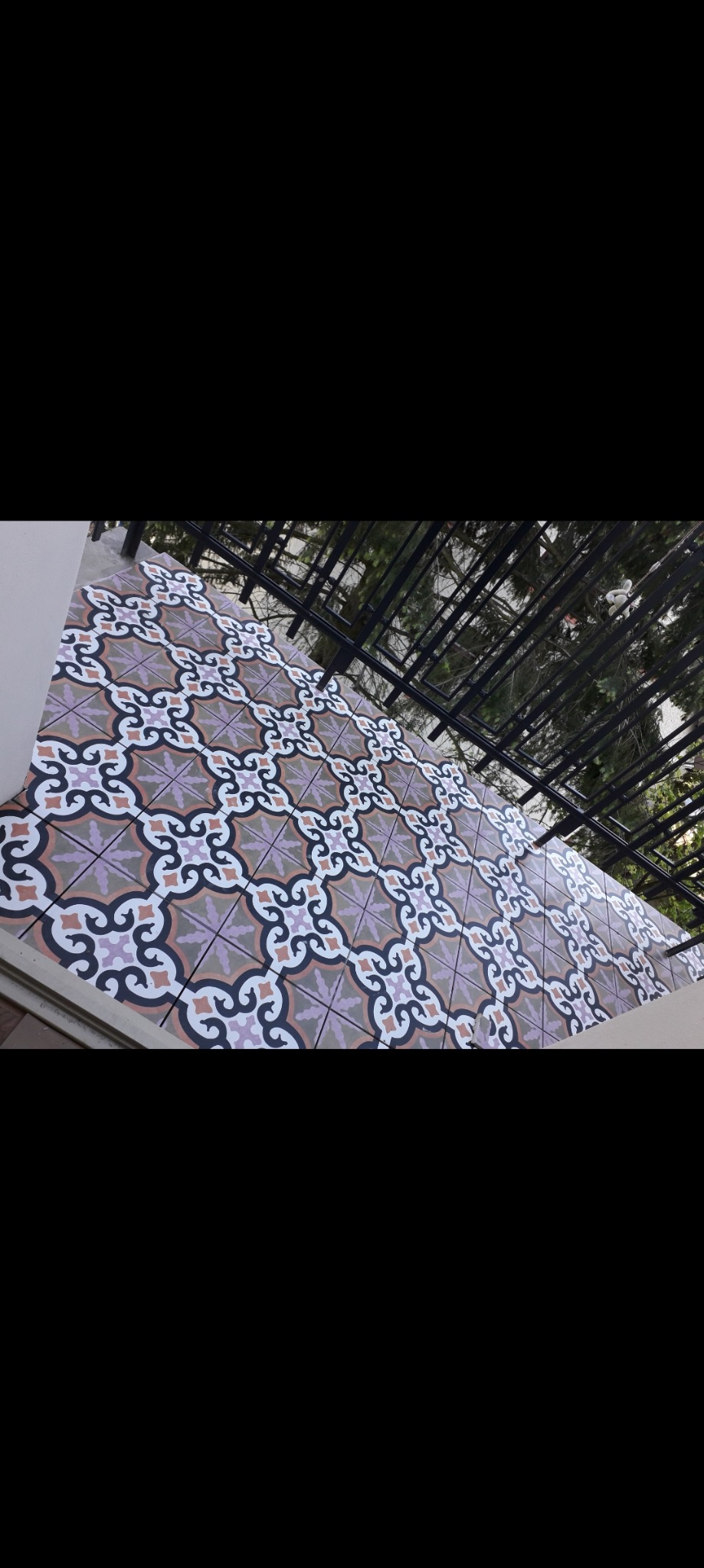 Balkon z dekoracyjną, wzorzystą terakotą w odcieniach fioletu, bieli i brązu. Widok z góry, z elementami konstrukcji zadaszenia w tle.