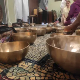 Uczestnicy sesji relaksacyjnej z misami dźwiękowymi na wzorzystym dywanie, w tle meble i osoby.