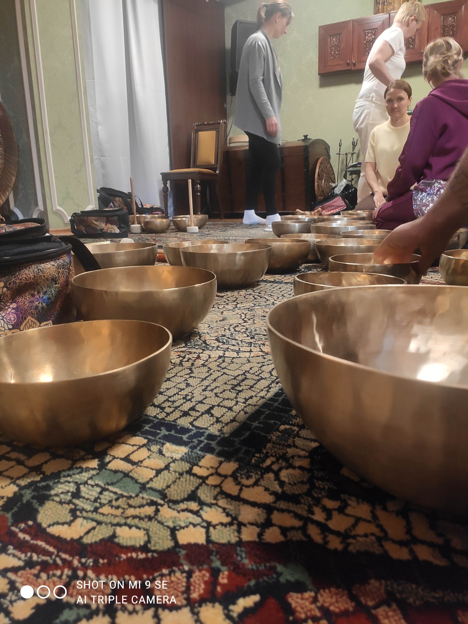 Uczestnicy sesji relaksacyjnej z misami dźwiękowymi na wzorzystym dywanie, w tle meble i osoby.