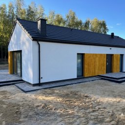 P. HOMES Sp. z o.o. - Nowoczesny, parterowy dom z białymi ścianami, czarnym dachem i drewnianymi akcentami, otoczony zielenią. Widoczne tarasy z kostki brukowej i fragment ogrodu.