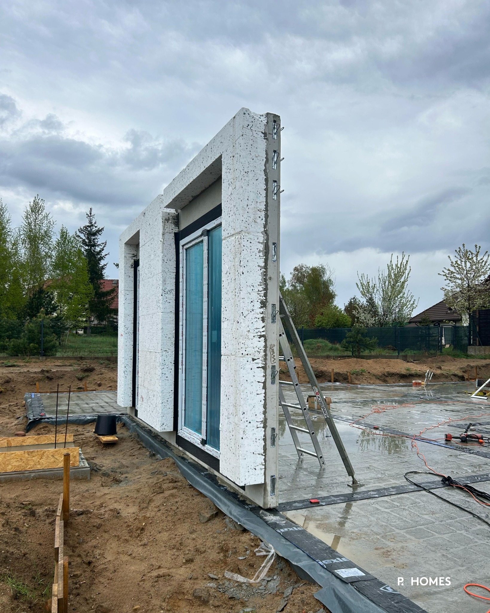 Montaż ściany domu z betonu komórkowego z oknem, widoczna drabina i fundament. Na zewnątrz pochmurne niebo, drzewa. Etap budowy.