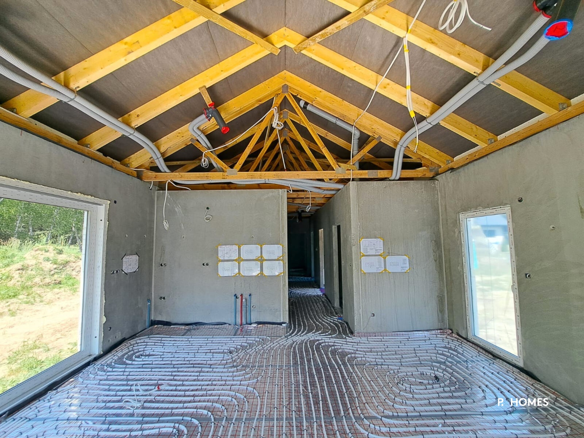 Wnętrze w surowym stanie z instalacją ogrzewania podłogowego, widoczne okna, drewniana konstrukcja dachu i instalacje elektryczne. Ściany w szarym kolorze.